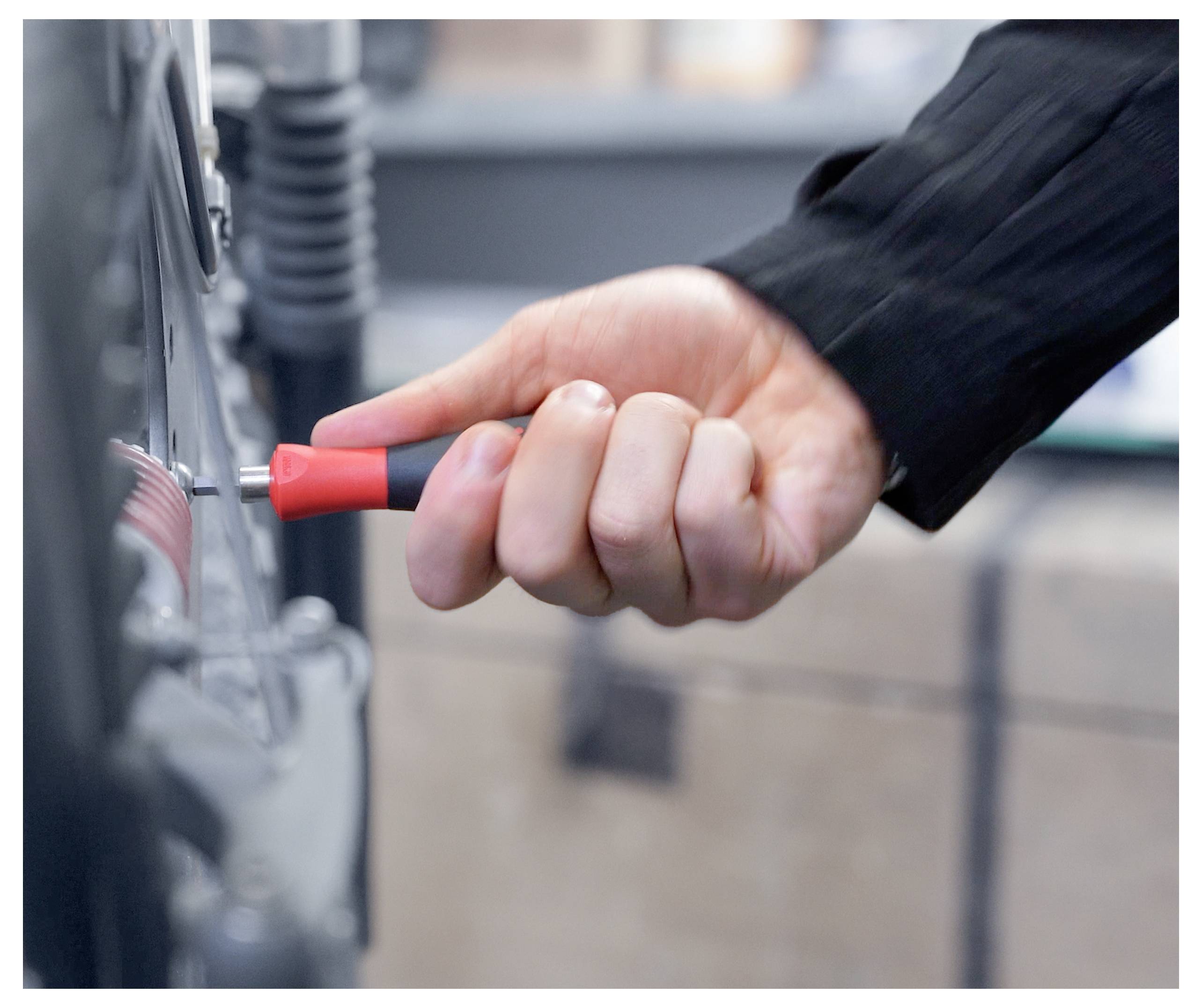 A hand in a black sleeve holds a screwdriver, tightening a bolt on machinery, focusing on precision work in an industrial setting.