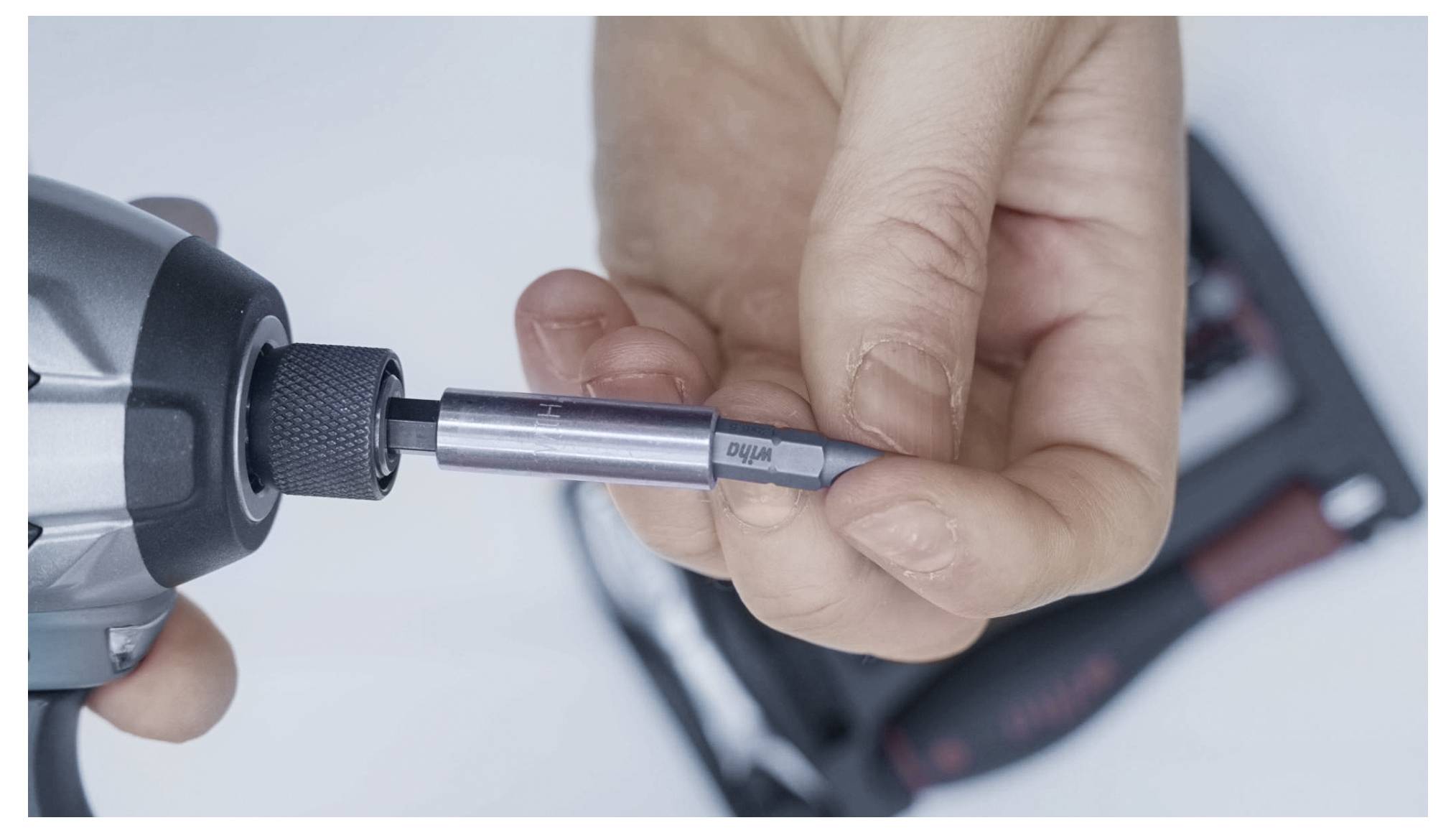A hand inserts a drill bit into an electric screwdriver, with blurred tools in the background.