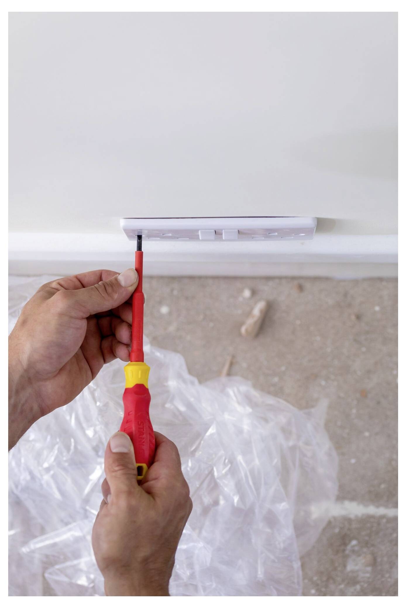 A person using a red screwdriver to install an electrical panel on a wall above a plastic sheet-covered floor.