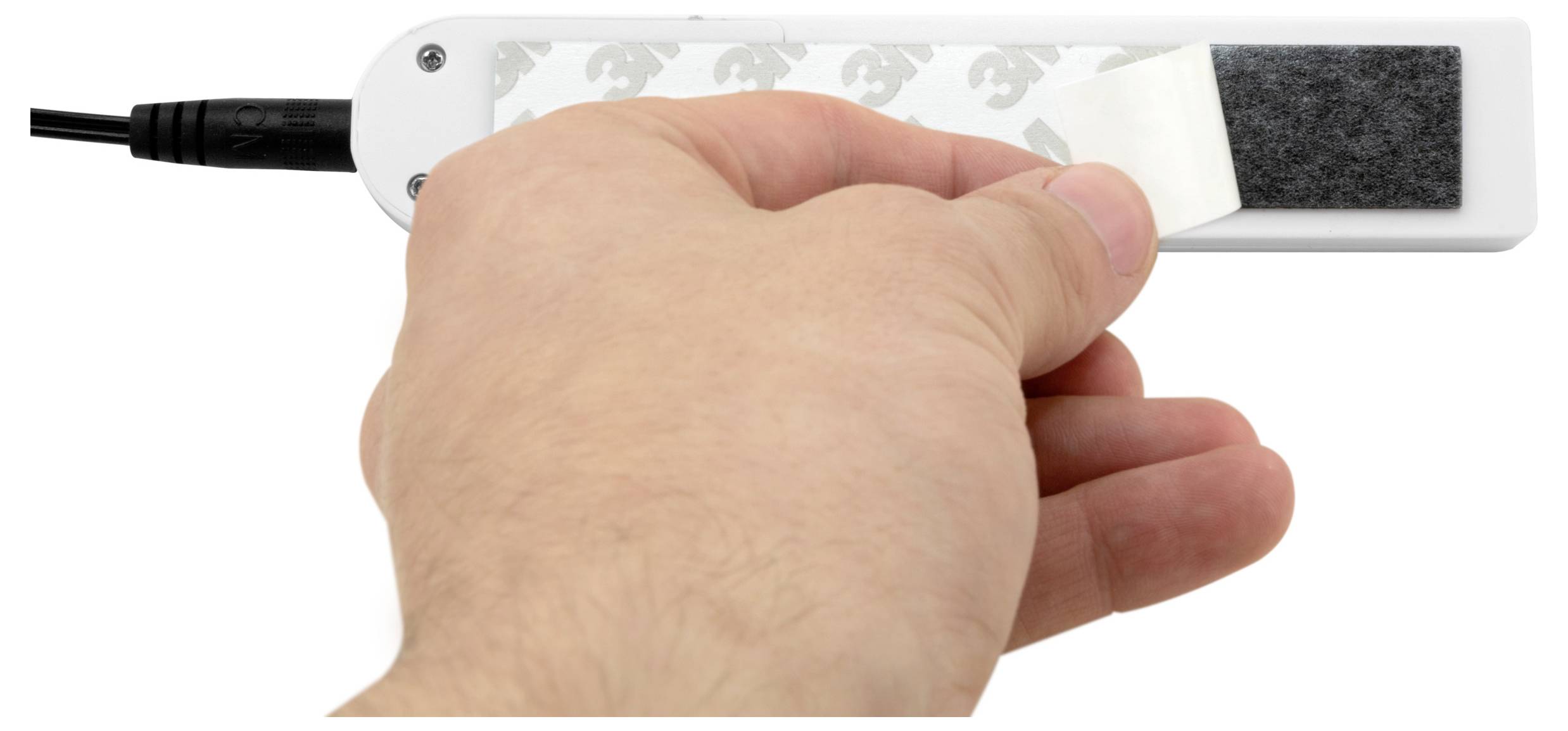 Hand peeling a white adhesive strip off a black-and-white rectangular device with a cable attached, likely removing a protective backing or mounting pad.