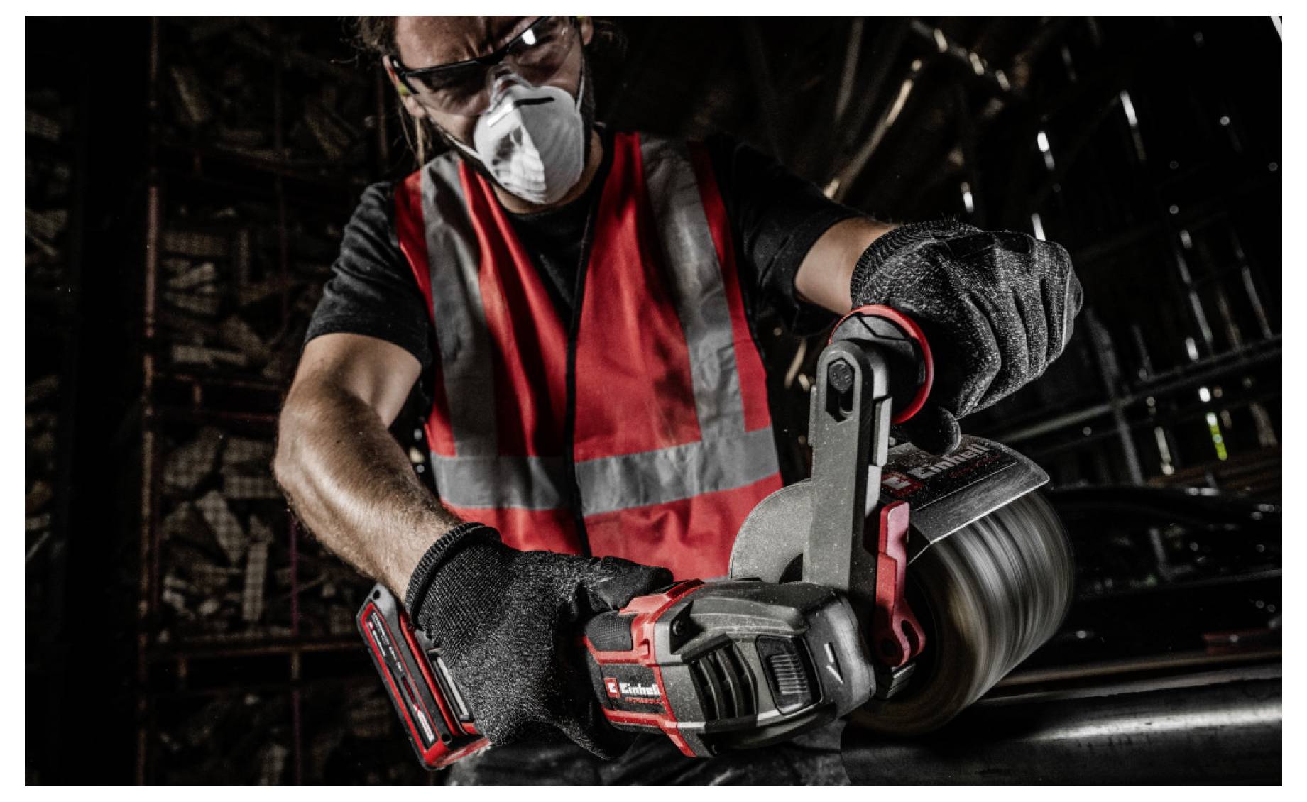 Worker wearing a mask and gloves uses a cordless pipe-cutting tool on a metal pipe in a dark industrial setting.