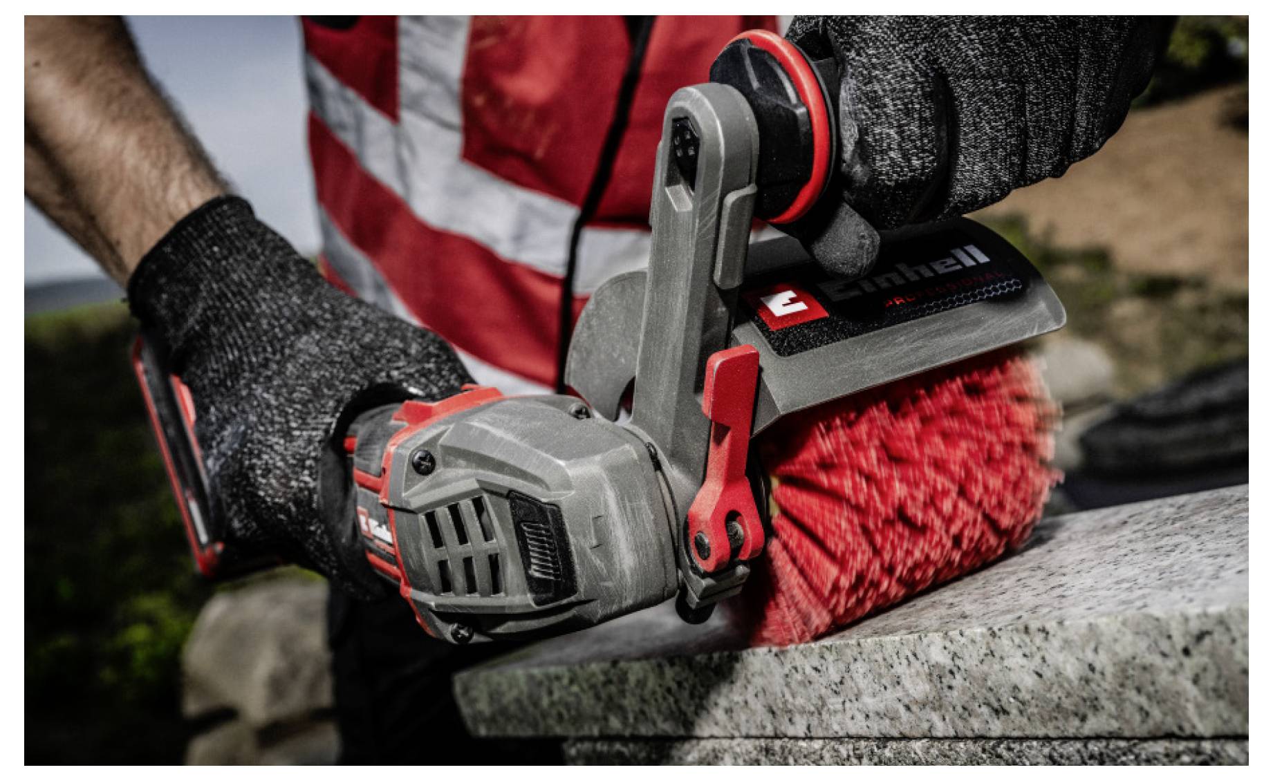 Gloved person using a handheld electric brush to clean a rough outdoor surface, with red bristles and gray-red tool visible close up.