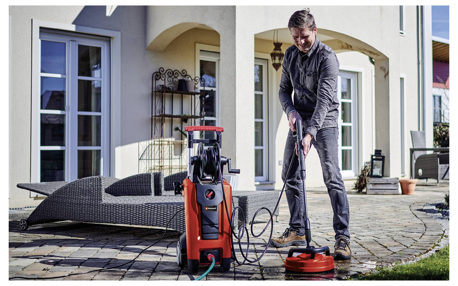 Man pressure-washes a patio with a red pressure washer and surface cleaner in front of a house.