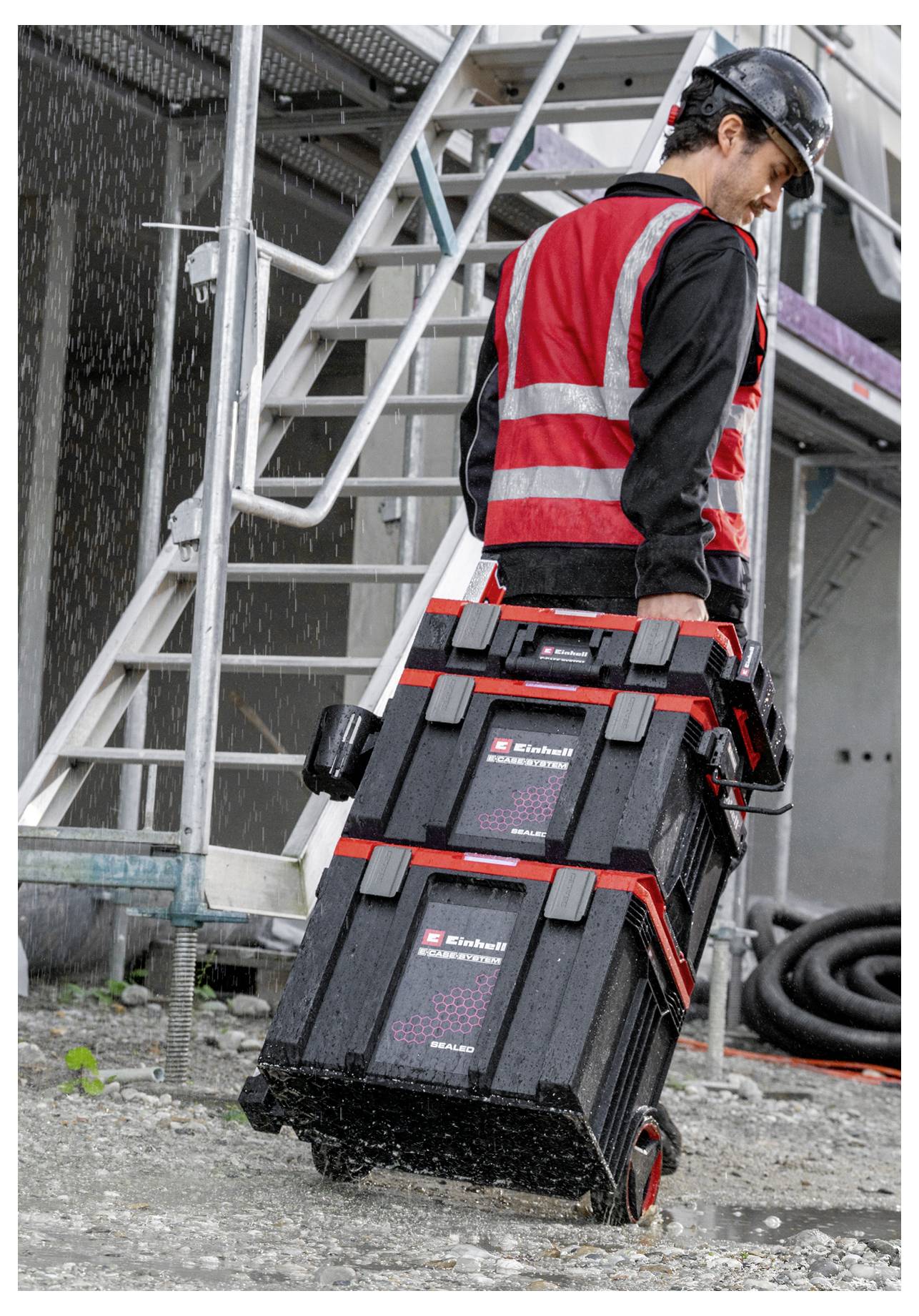 Construction worker in a hard hat and high-visibility vest carries two stacked tool cases at an industrial site beside metal stairs.