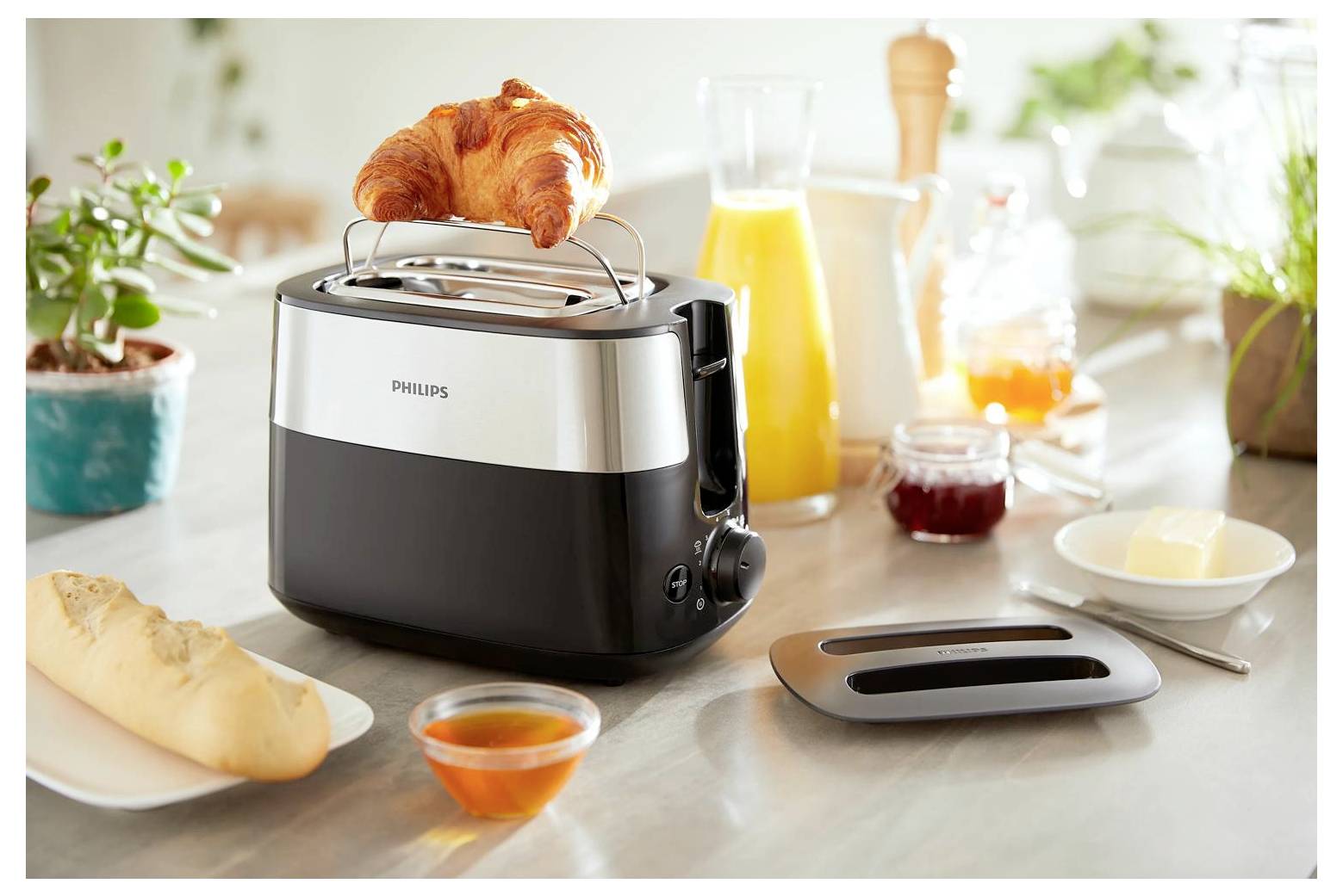 Croissant in a toaster on a kitchen counter with bread, juice, jam, butter, and a knife, suggesting a breakfast scene.