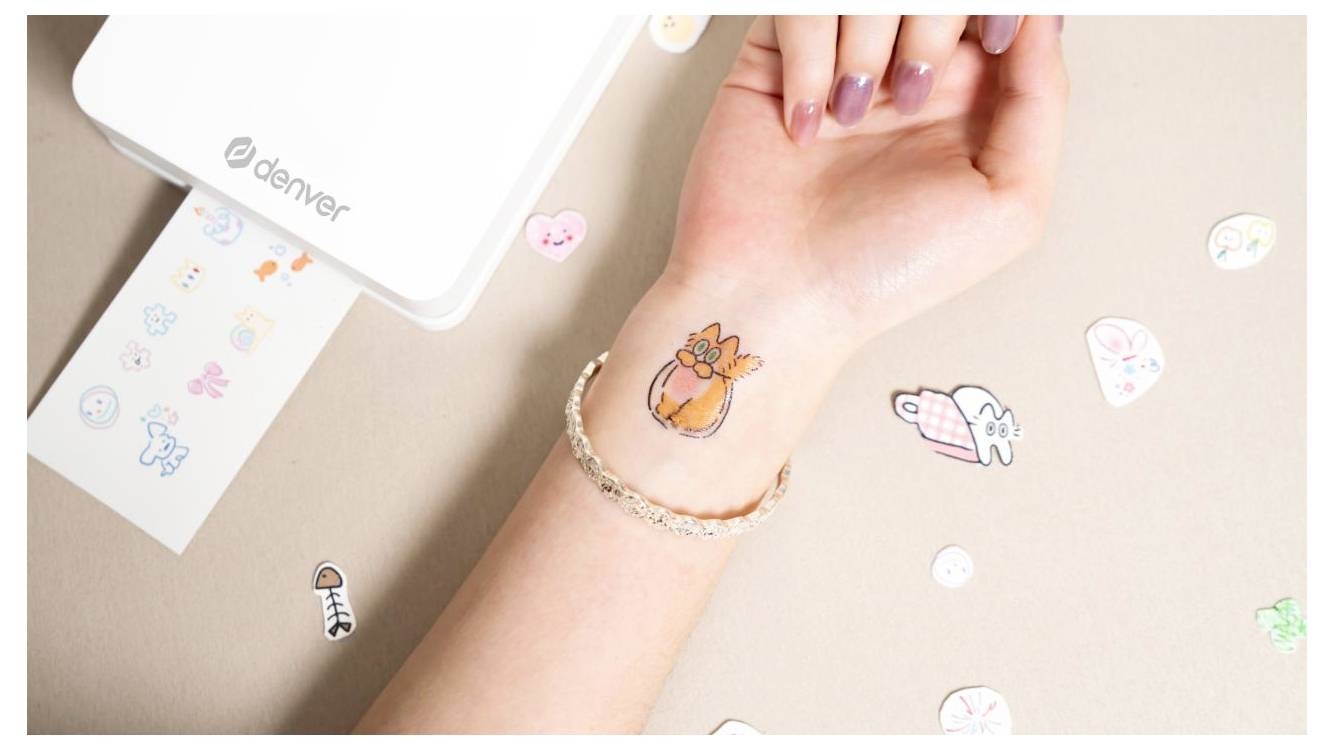 Hand applying a small orange flower temporary tattoo to a wrist beside a sticker printer and scattered decorative stickers.