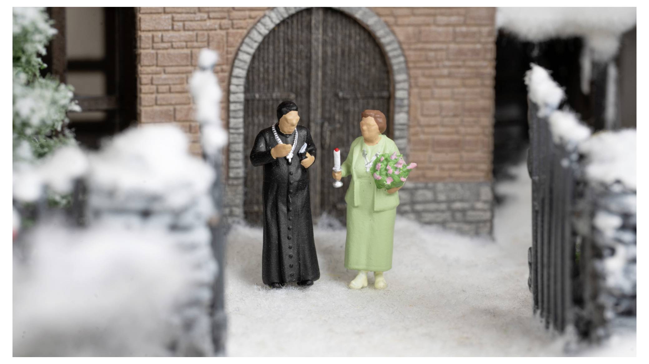 Miniature bride and groom figurines stand outside a brick building, dressed in black and green and holding flowers and a candle.