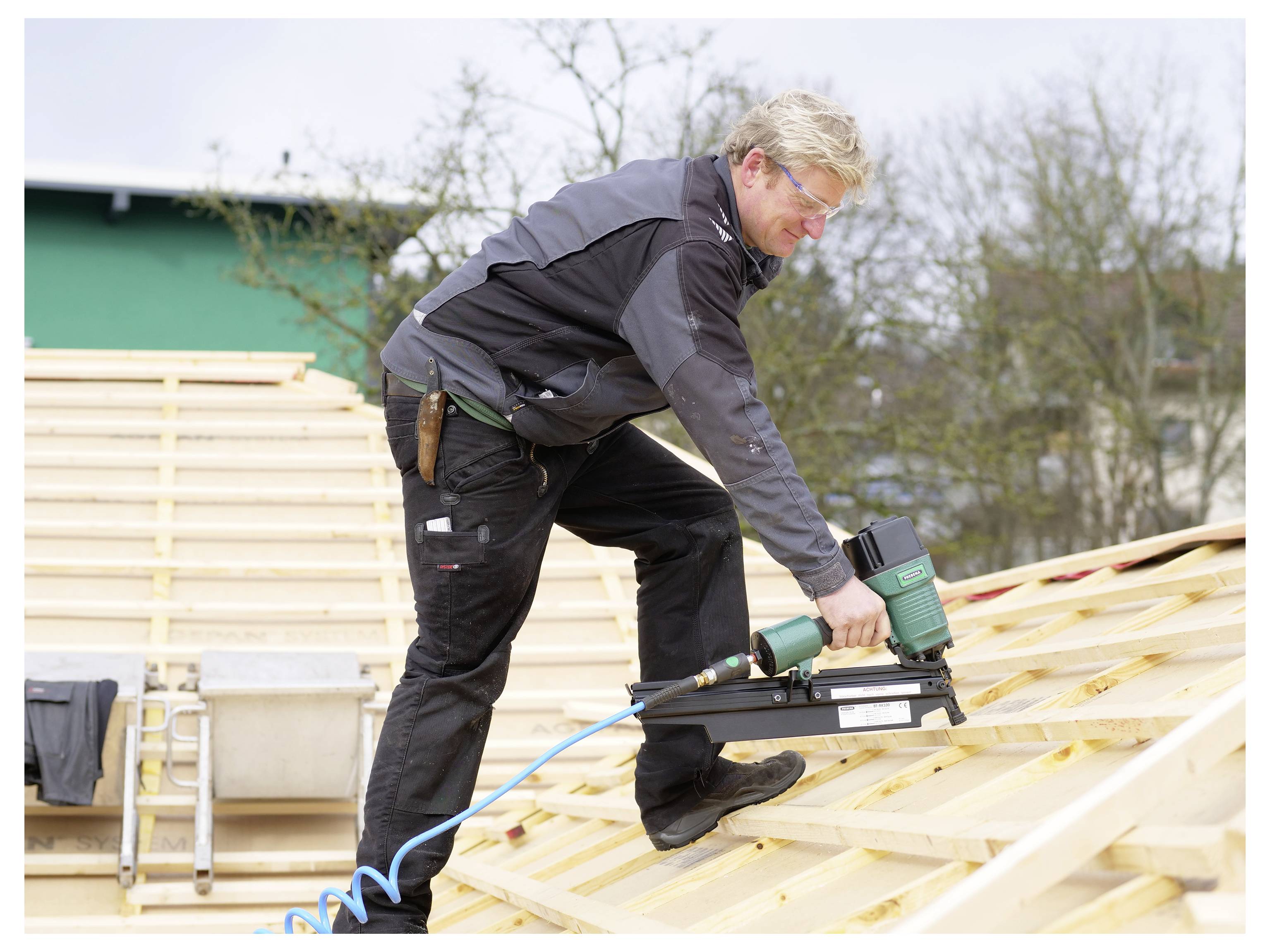 Worker using a nail gun to fasten wooden roof trusses outdoors on a construction site.