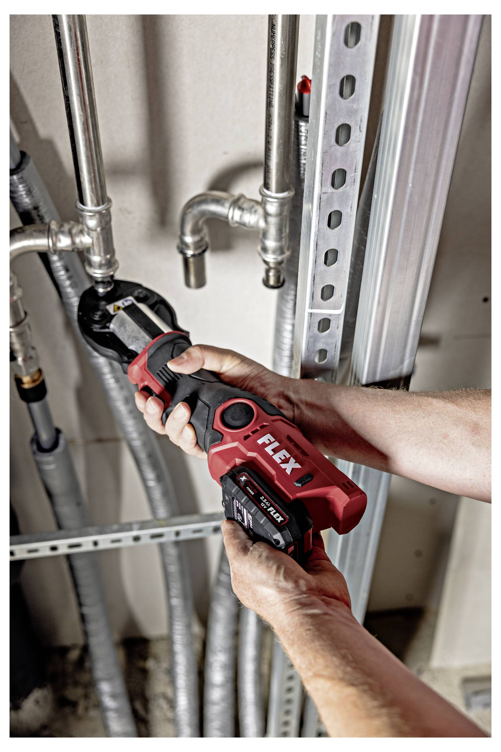 Person uses a red FLEX cordless pipe cutter on metal plumbing pipes inside a utility space.