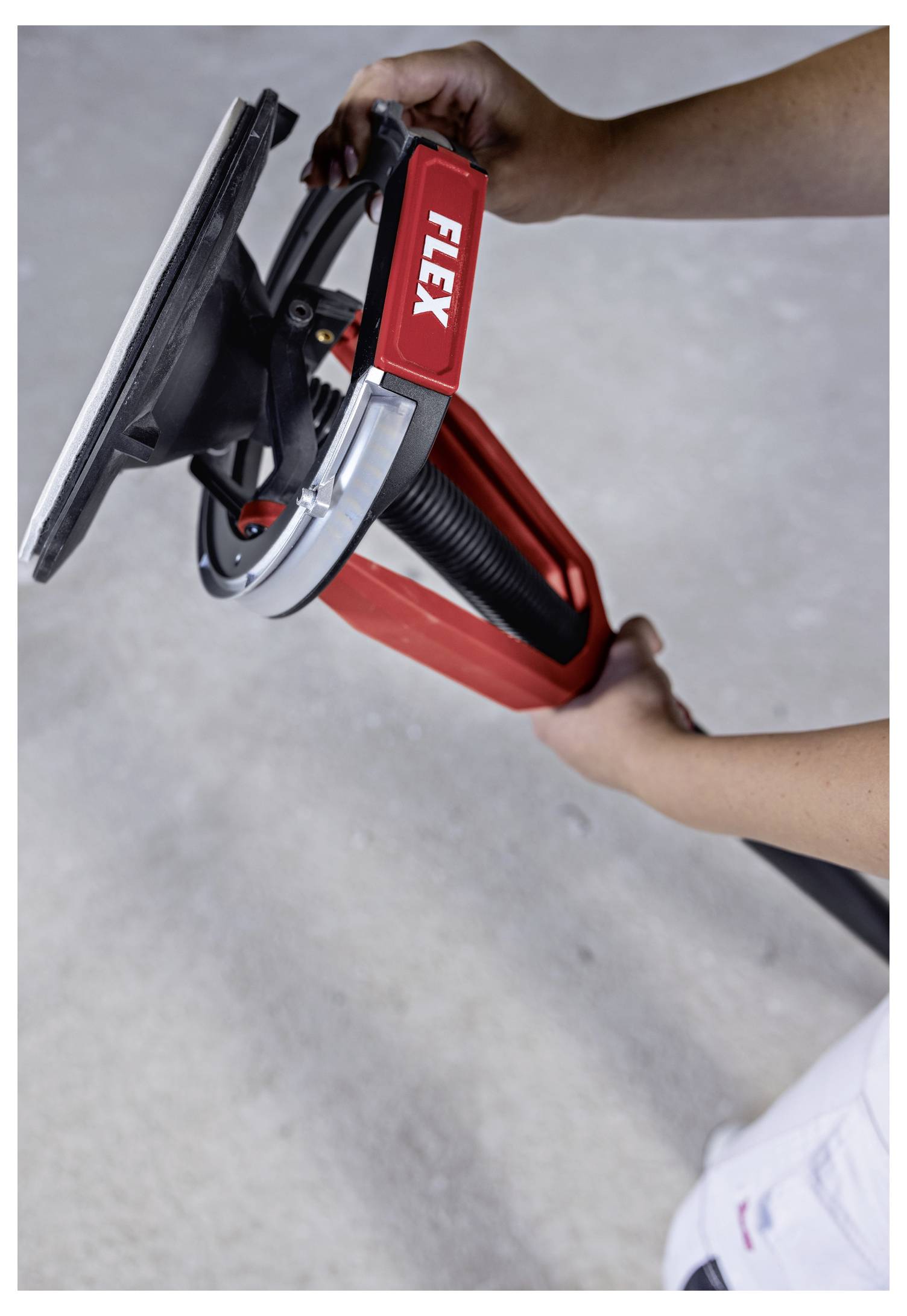 Hands holding a red FLEX power sander or polisher with a rectangular pad, shown in close-up against a plain gray background.