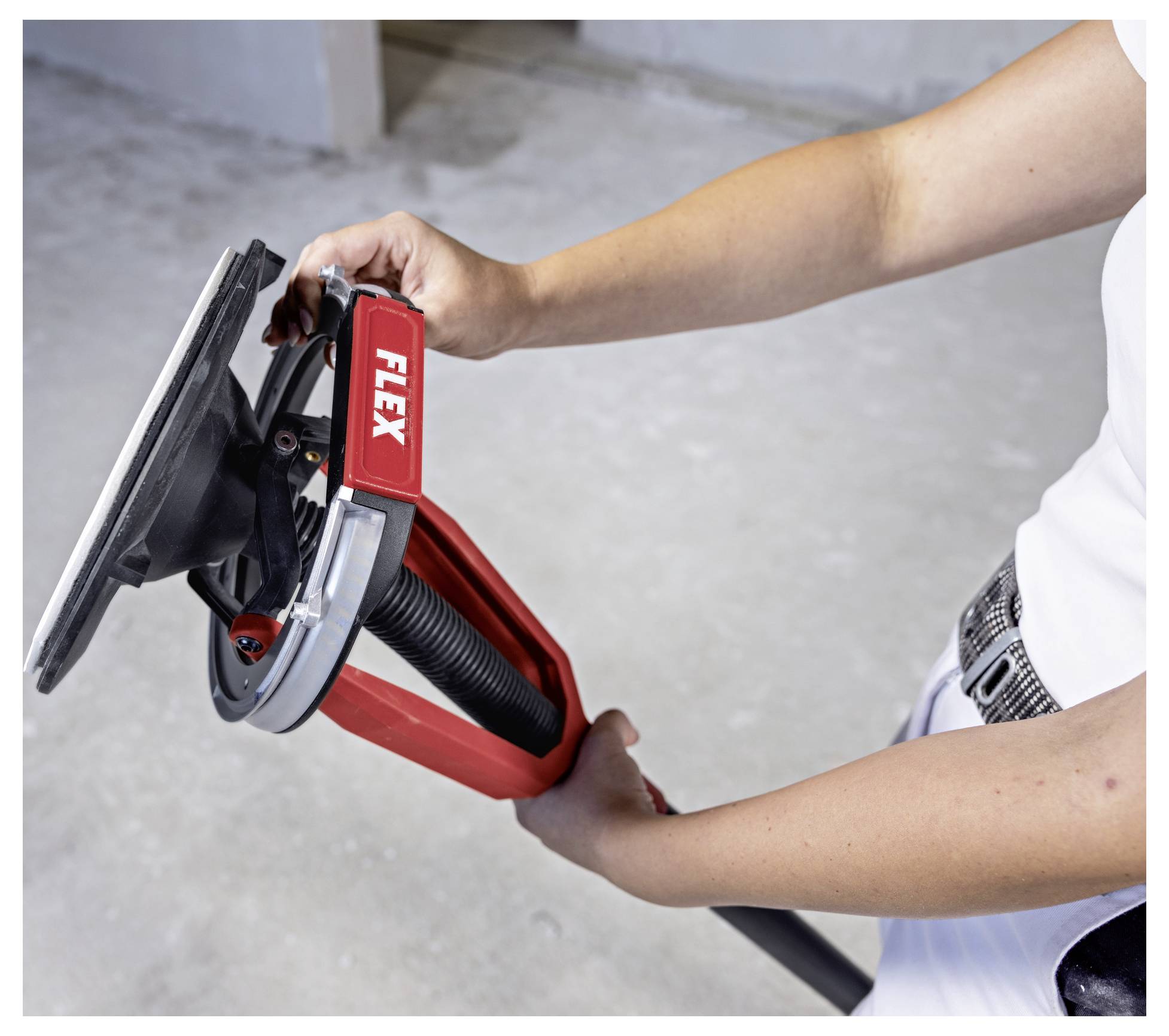 Person holding a red FLEX drywall sander with a round sanding head and vacuum hose attached, shown in use on a wall surface.