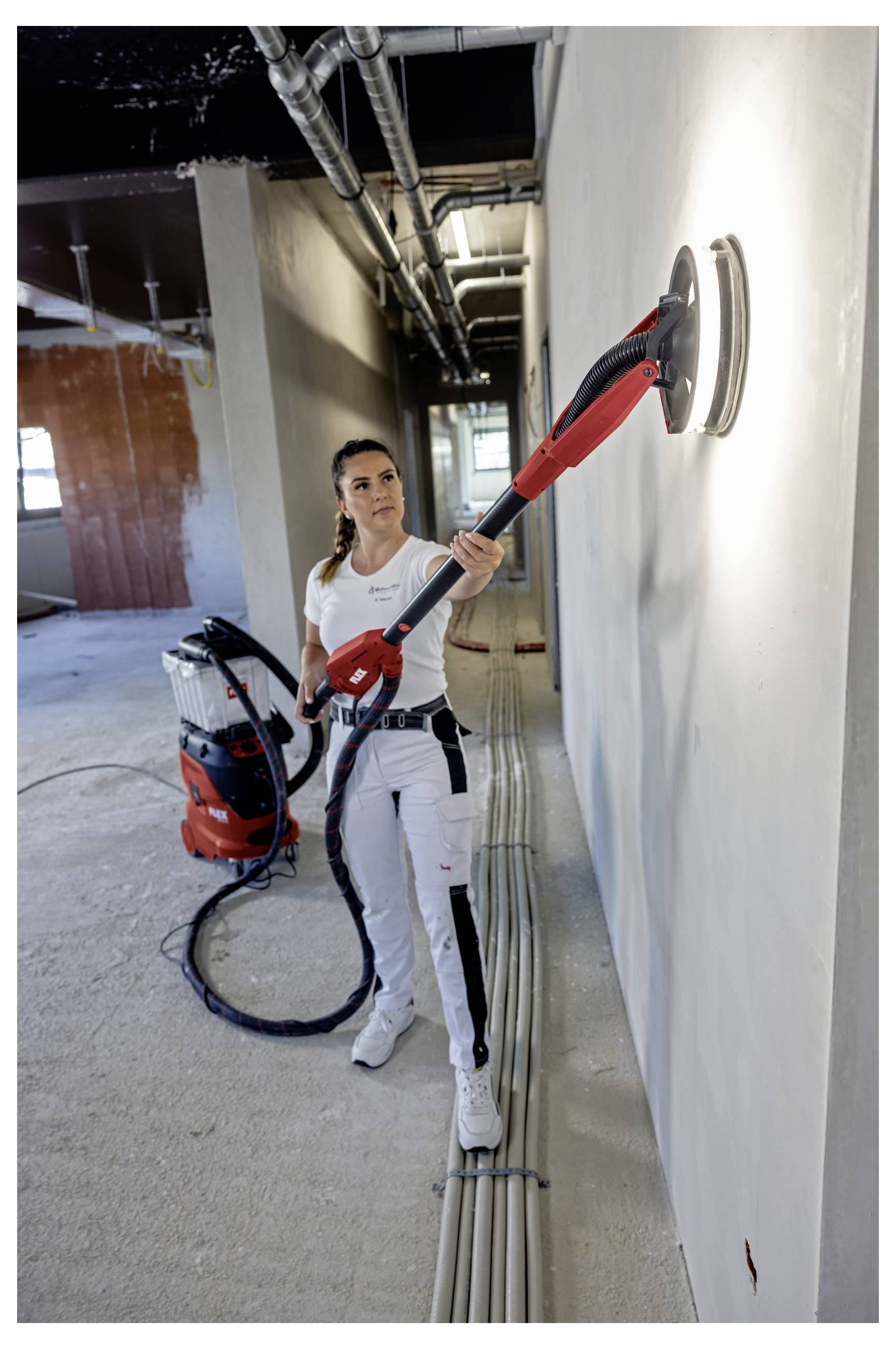 A worker sands a wall with a long-handled tool in an unfinished building corridor, with a vacuum system on the floor behind her.