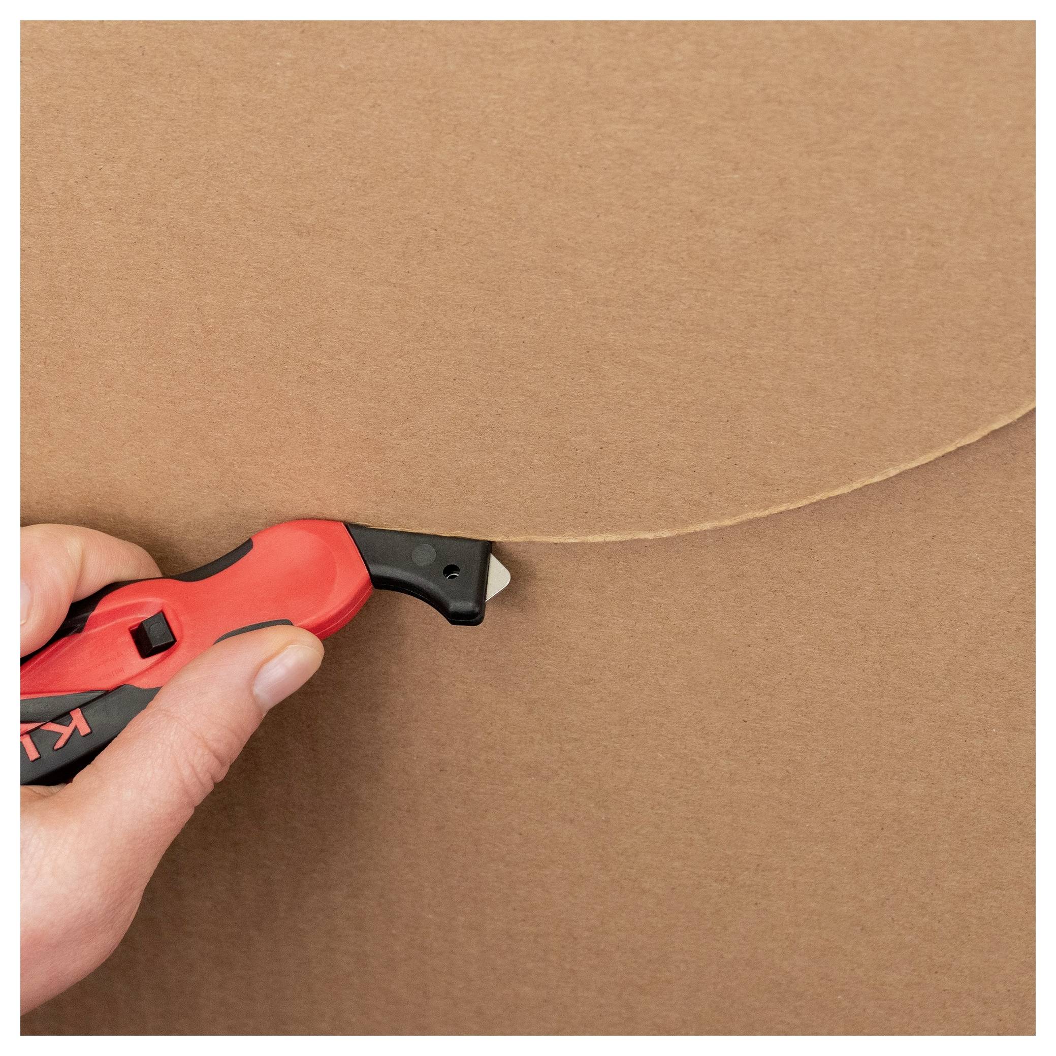 Hand using a red utility knife to cut a curved line into brown cardboard.