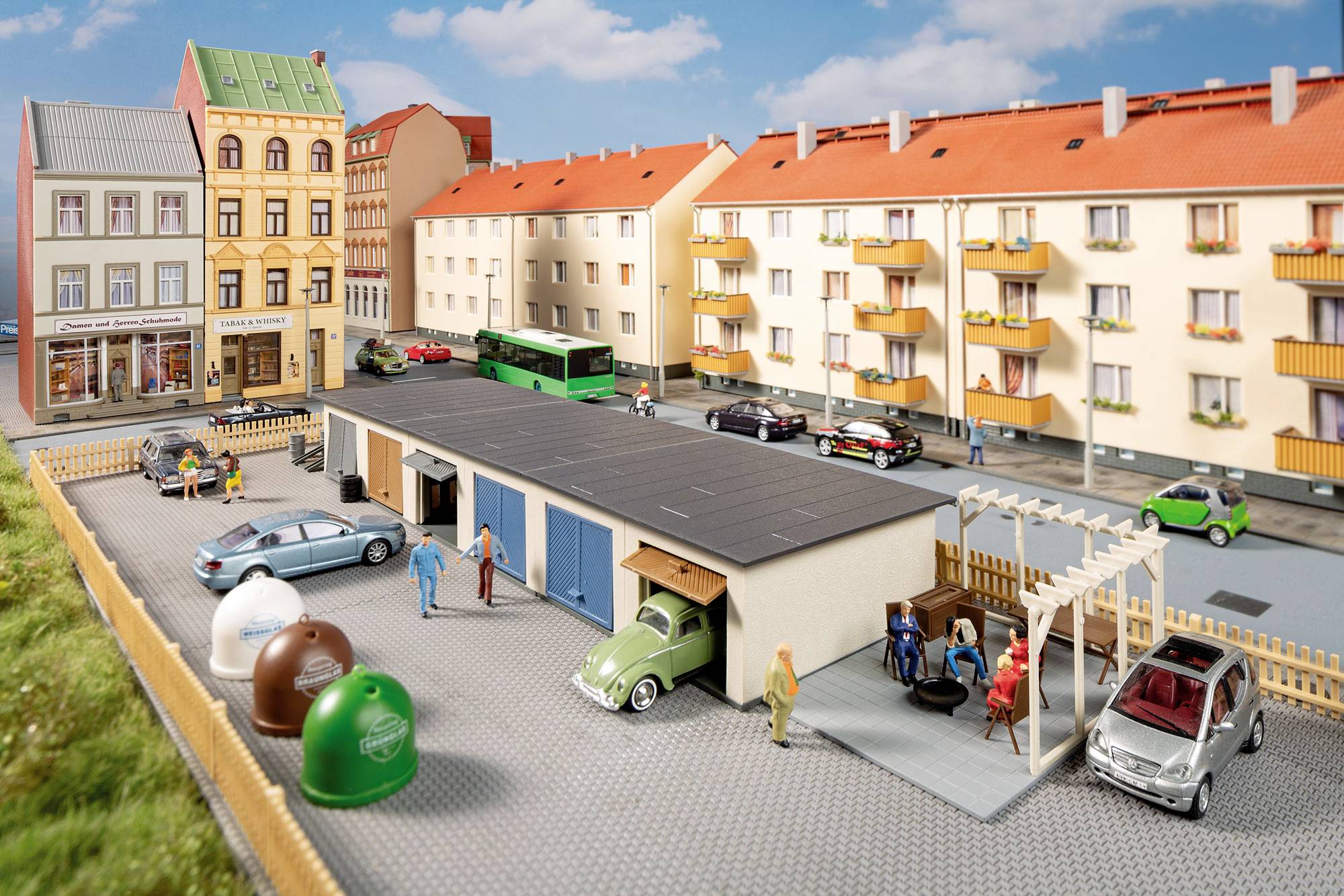 Miniature street scene with apartment buildings, parked cars, pedestrians, a green bus, and a small yard with recycling bins and a garage