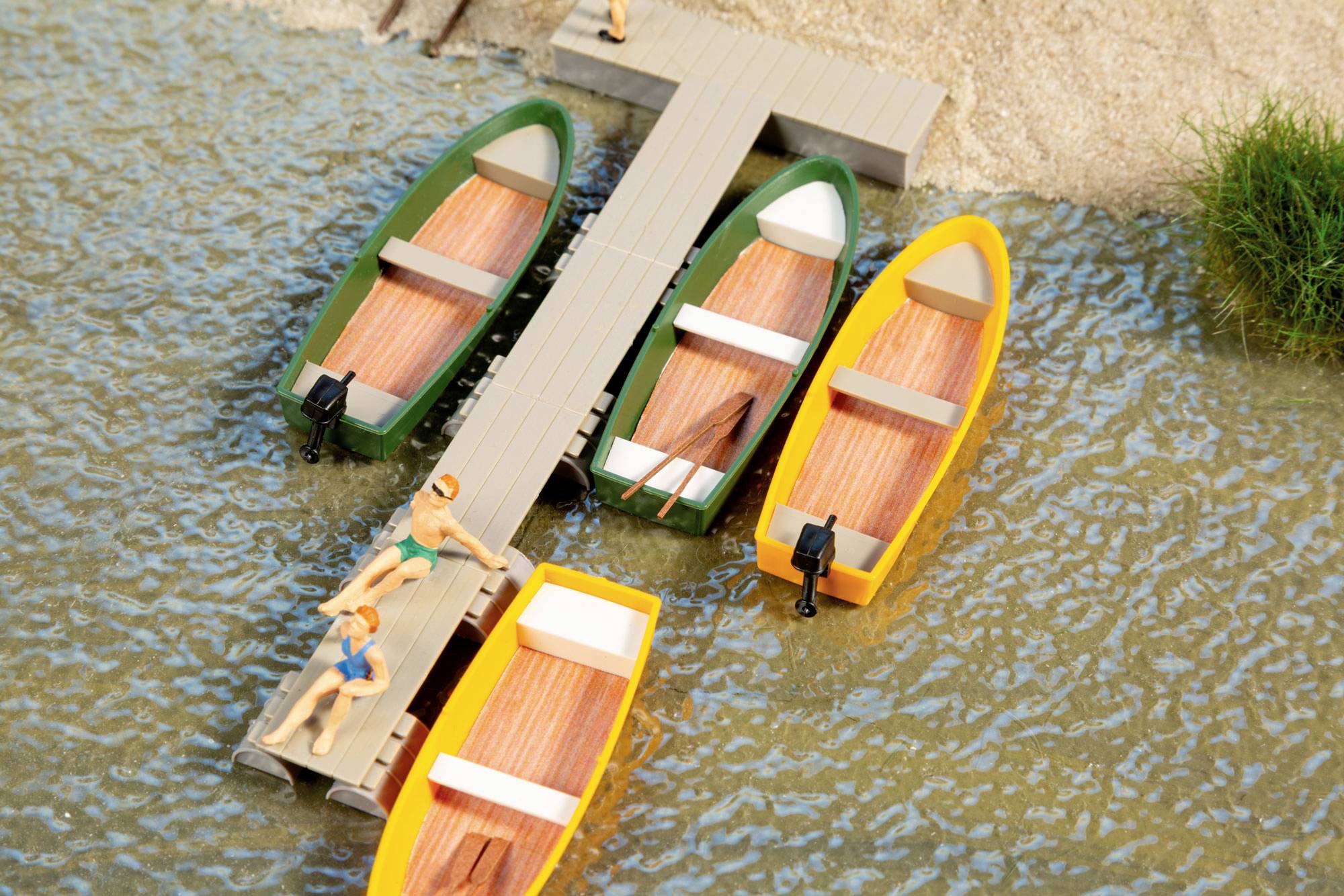 Miniature dock with four colorful boats moored in calm water, with two small figures on the pier near the shore.