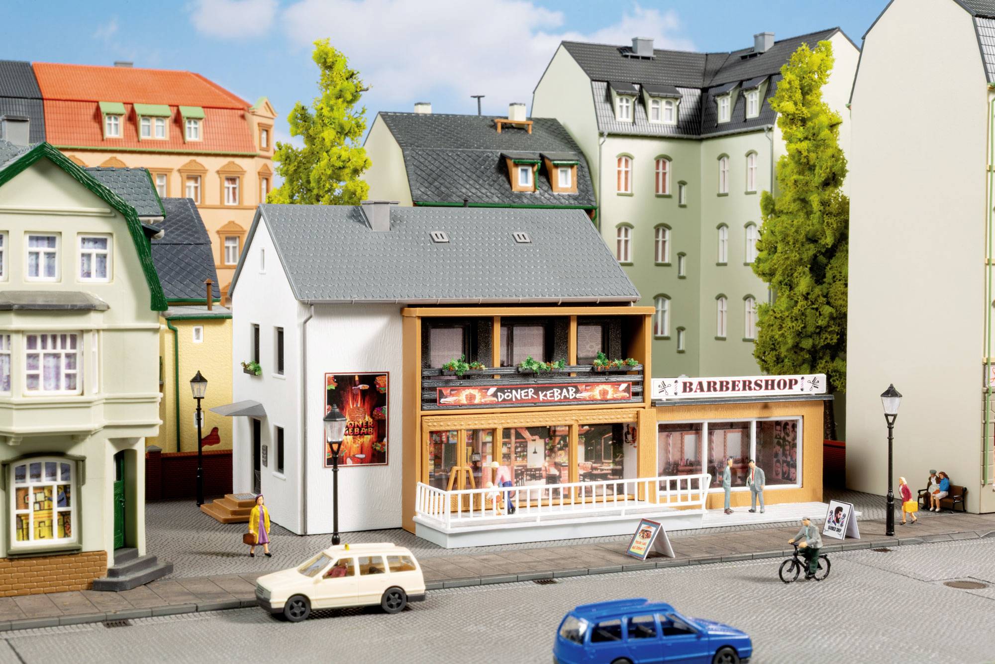 Miniature street scene with a kebab restaurant and barbershop on a row of model buildings, with tiny cars, pedestrians, and a cyclist.