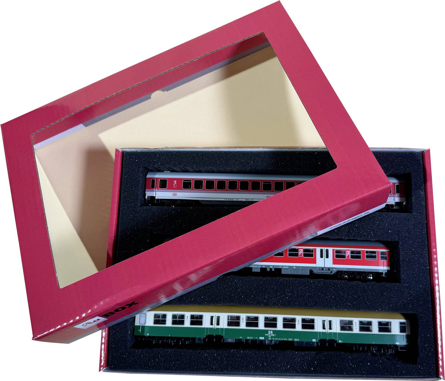 Boxed model train set with three miniature passenger cars in a foam tray, partially covered by a red lid with a clear window.