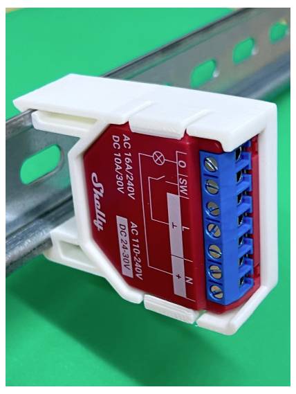 Close-up of a Shelly electrical relay module mounted on a DIN rail, with blue screw terminals and labeled AC/DC wiring connections.