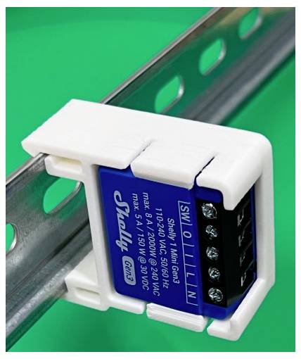 Close-up of a Shelly 1 Mini Gen3 smart relay module mounted on a metal DIN rail against a green background.