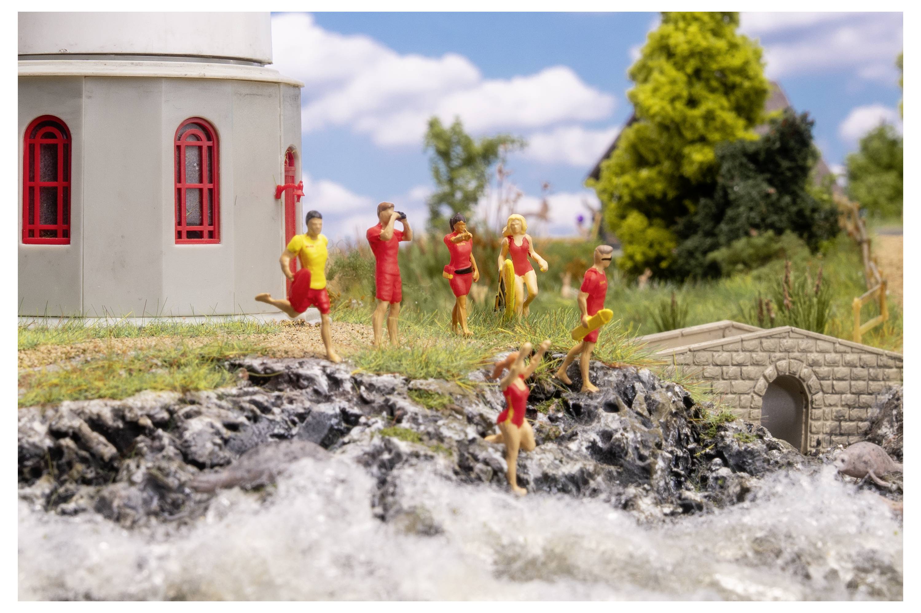Miniature scene of people in red and yellow clothing standing and running on rocky ground beside a small stone bridge and church-like building near water.