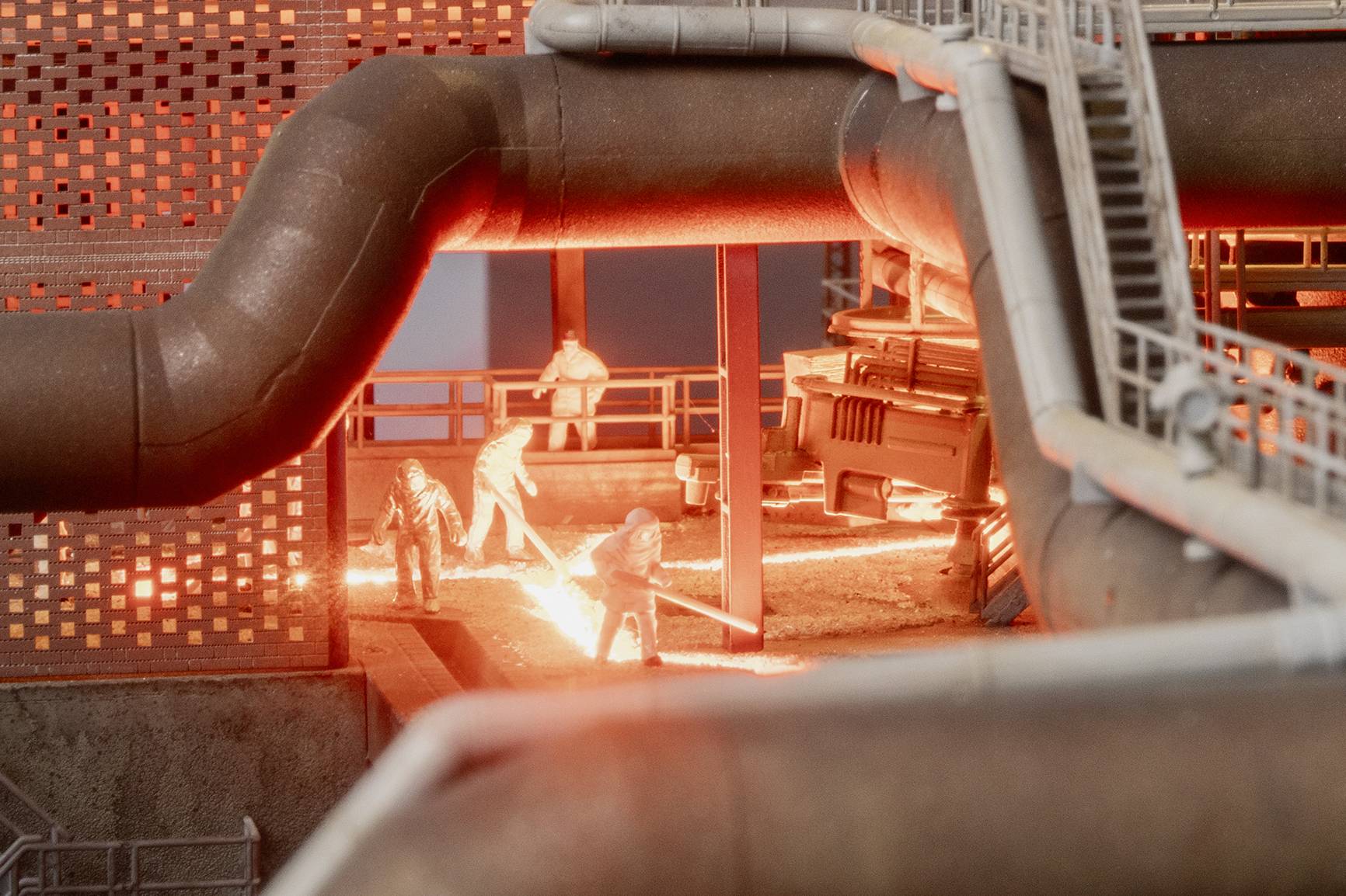 Miniature industrial scene with workers in protective gear around bright molten metal or welding sparks beneath large pipes and a metal structure.