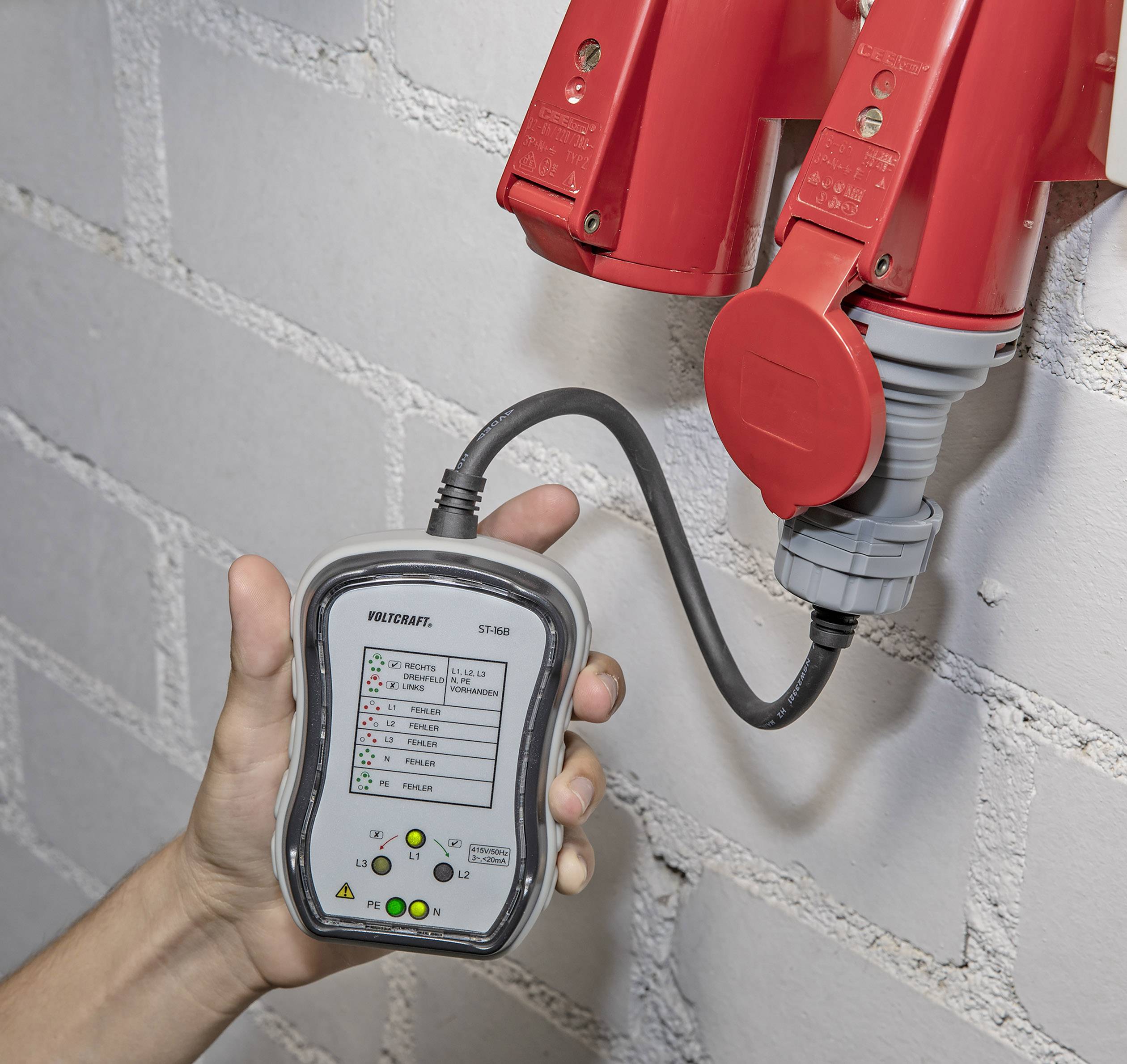 A hand is holding a testing device connected to red industrial sockets to check electrical voltage and phase configuration.