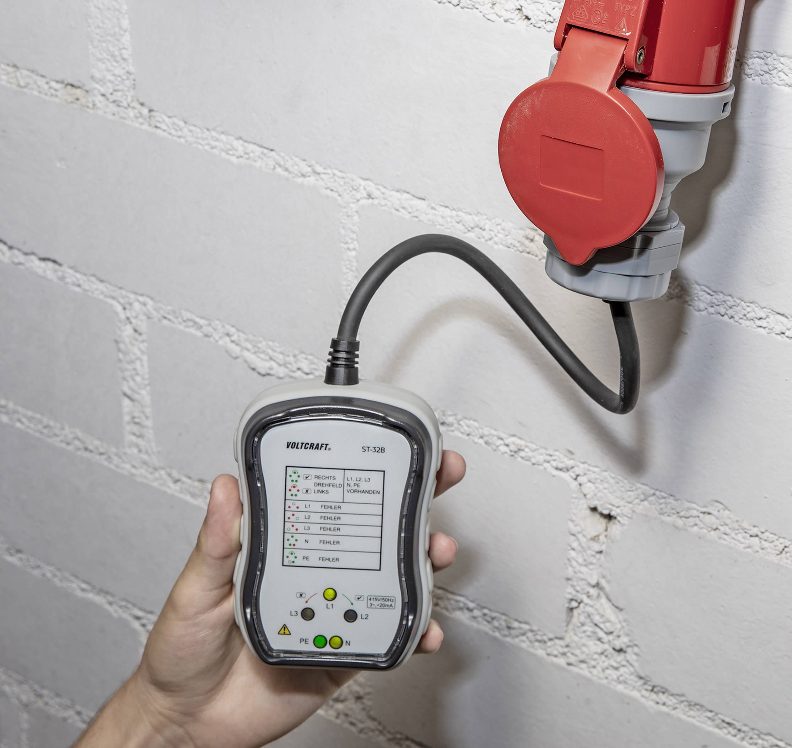 A hand is holding a device for checking electrical connections. It is connected to a red socket on a brick wall.