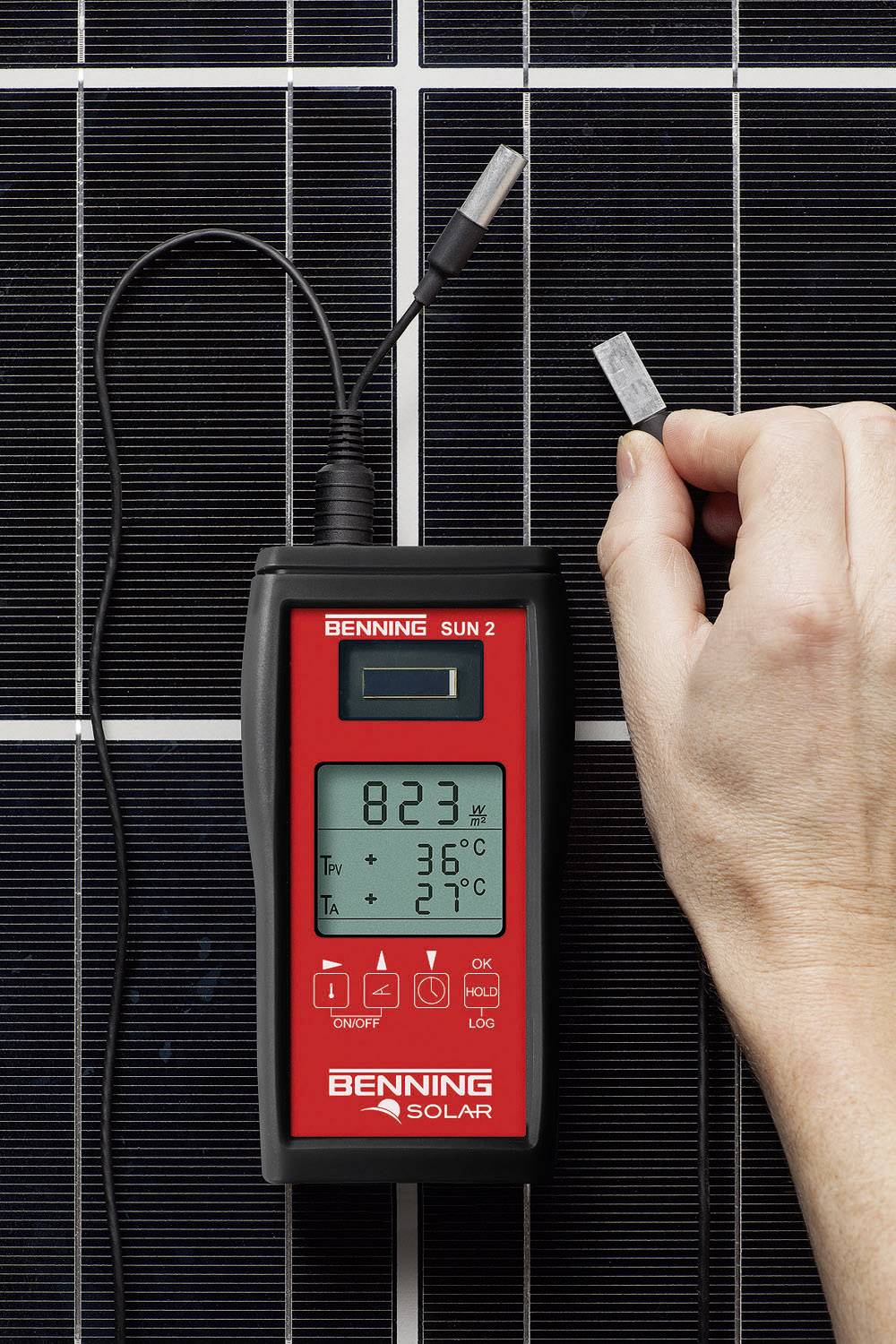 A hand holds a thermometer in front of a solar panel, showing a temperature of 36°C with an ambient temperature of 27°C.