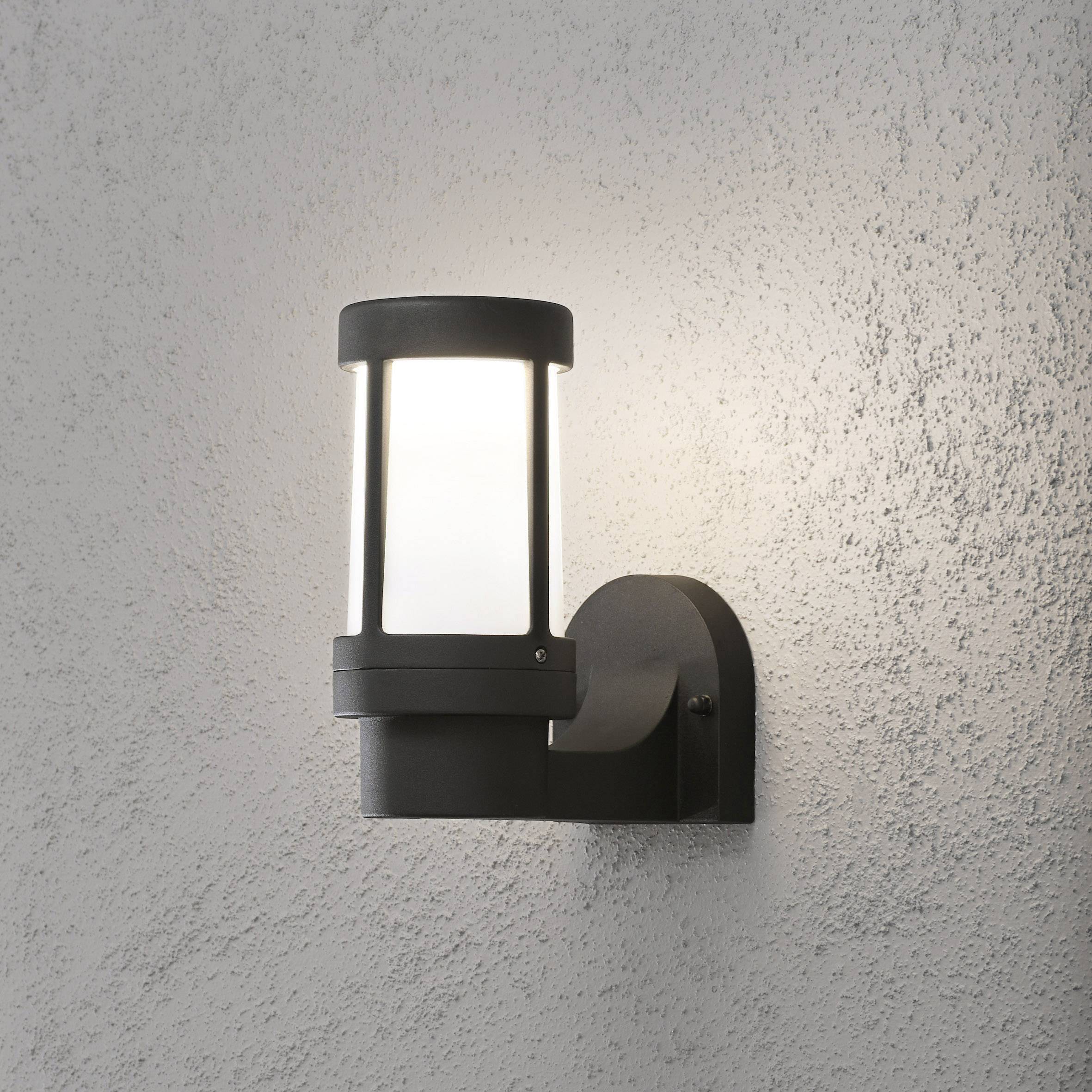 A contemporary external wall light made of black metal with white glass, mounted on a grey wall, emits soft light.