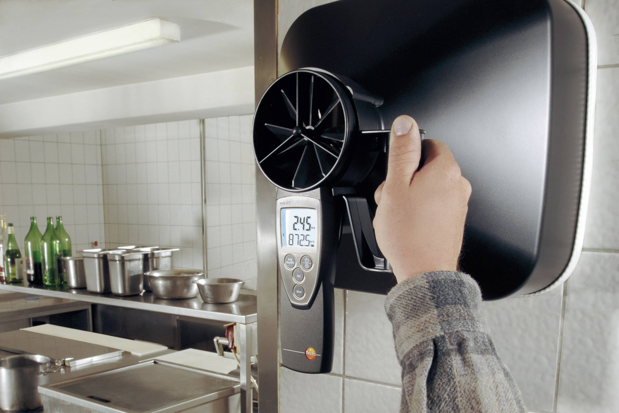 A handheld device for measuring air flow is used in an industrial kitchen. A display shows the measurement data.