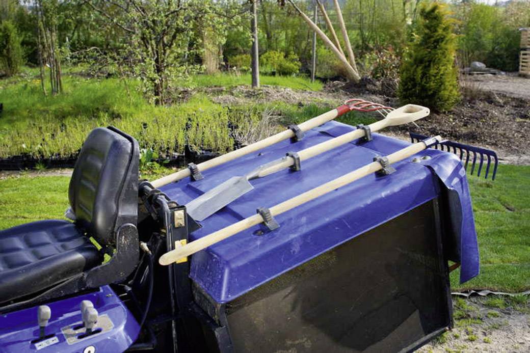 Lawn tractor with attached gardening tools such as a spade, rake, and shovel. In the background, a garden with trees and shrubs.