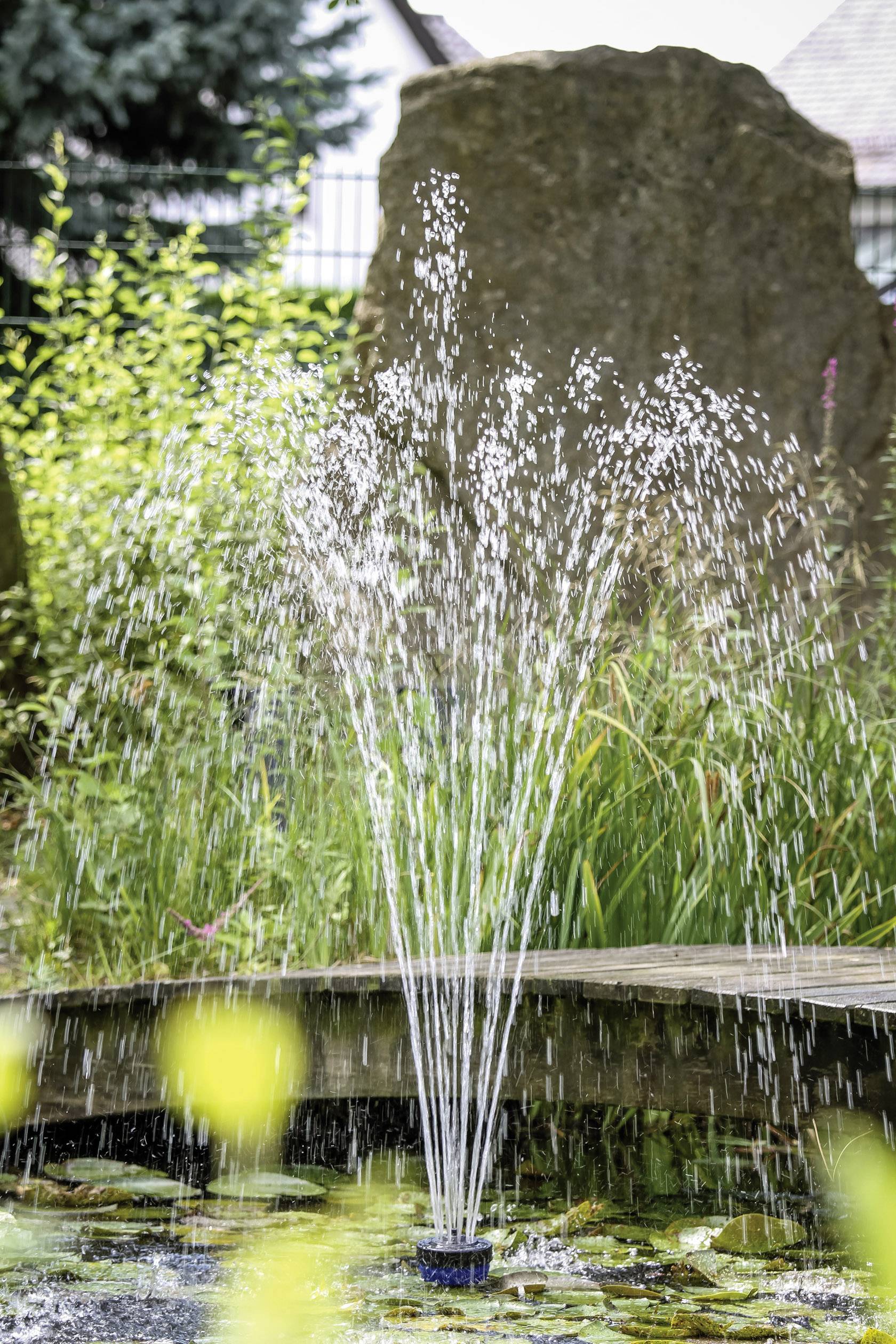 Springbrunnen im Garten mit hohem Wasserstrahl, umgeben von Pflanzen und Felsen.