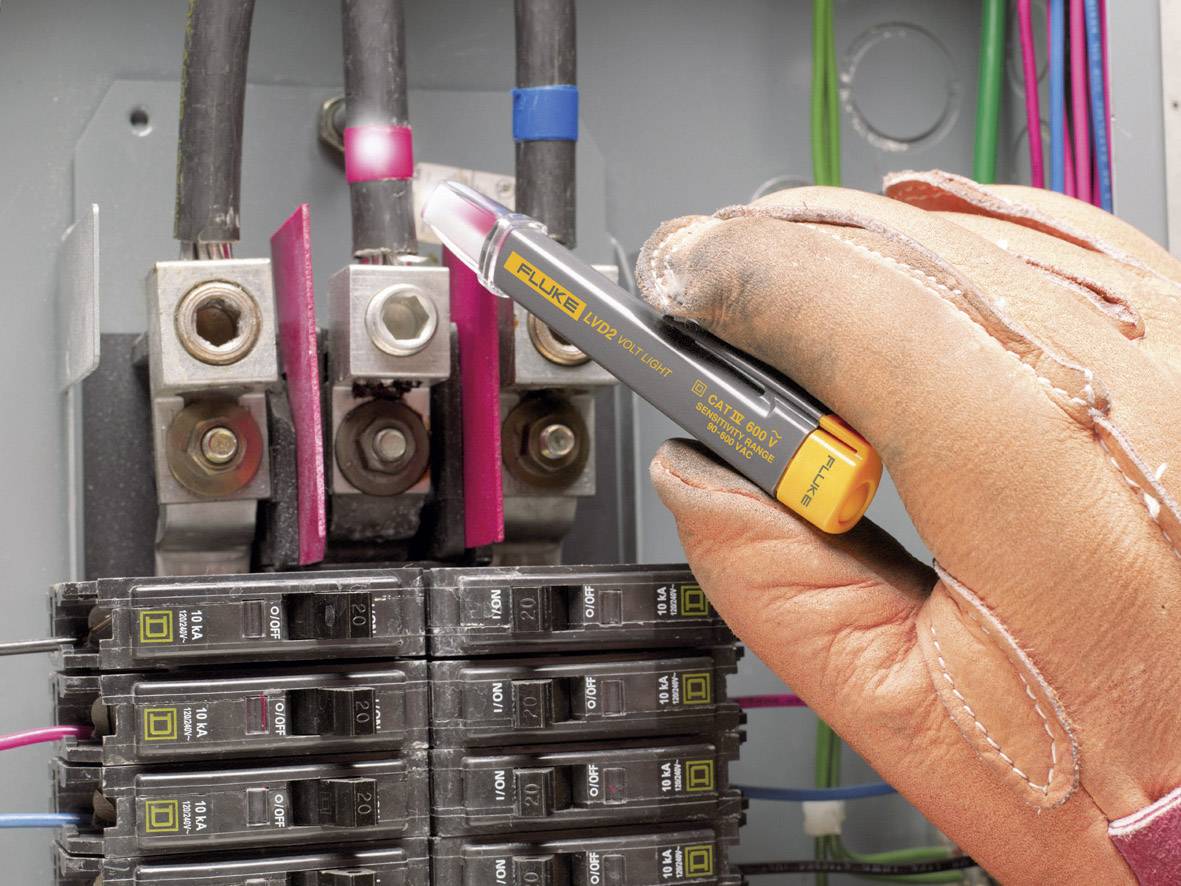 A hand wearing a glove is holding a voltage tester on an electrical circuit. Fuses and cables are visible. Safety inspection.