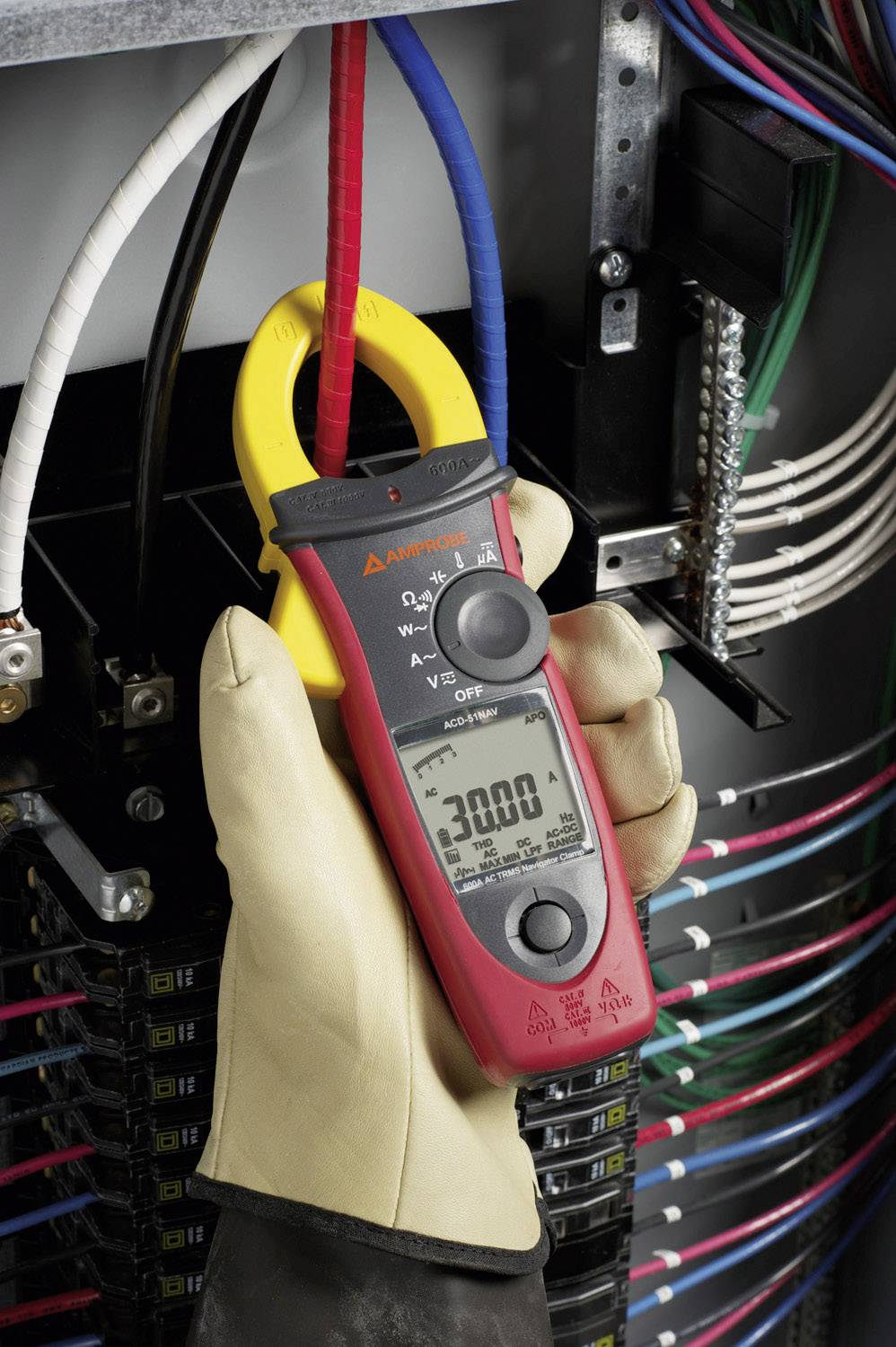 A gloved hand holds an amperemeter clamp around a red cable conductor. The display shows '3000'. Numerous coloured cables are visible in the background.
