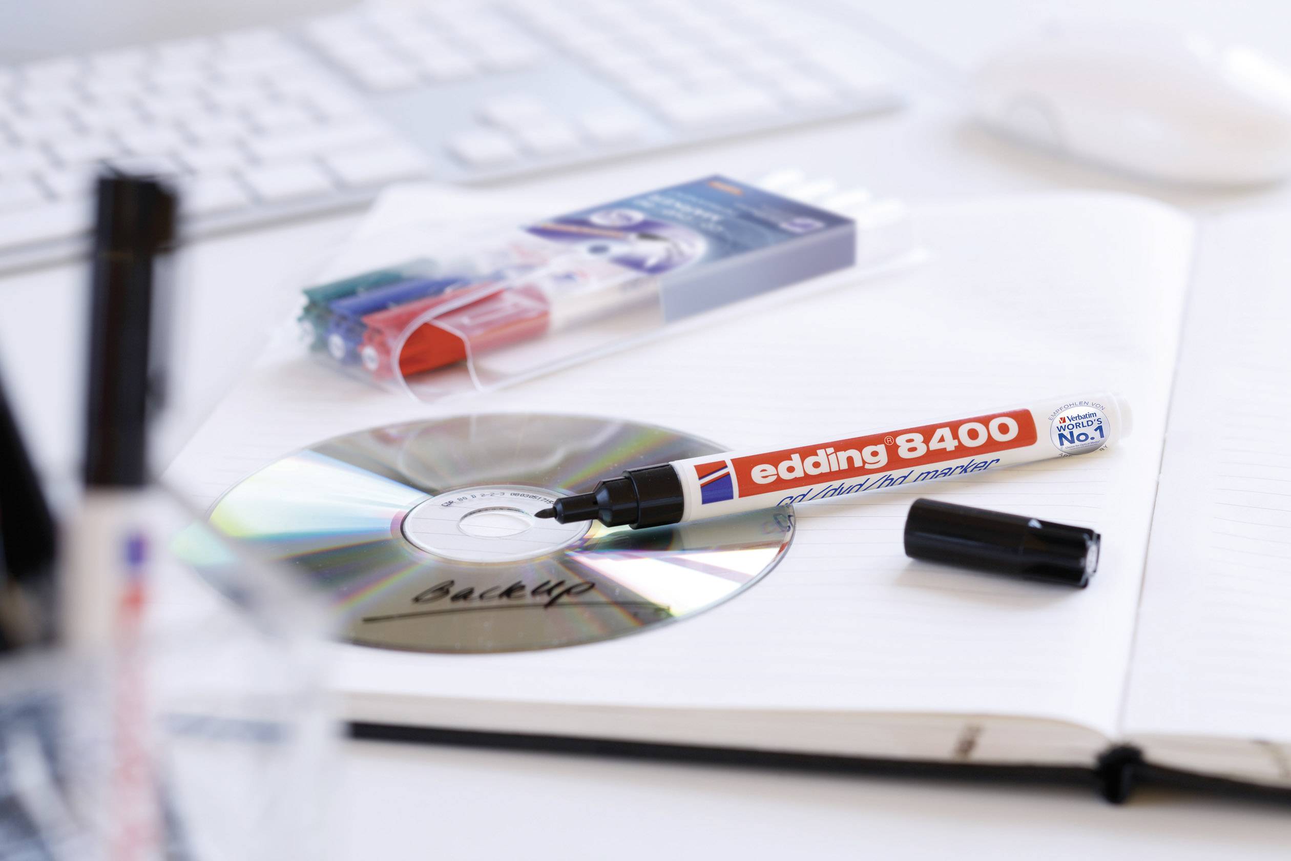 A red CD marker is lying on a DVD and a notebook, next to an opened stationery set and a computer keyboard.