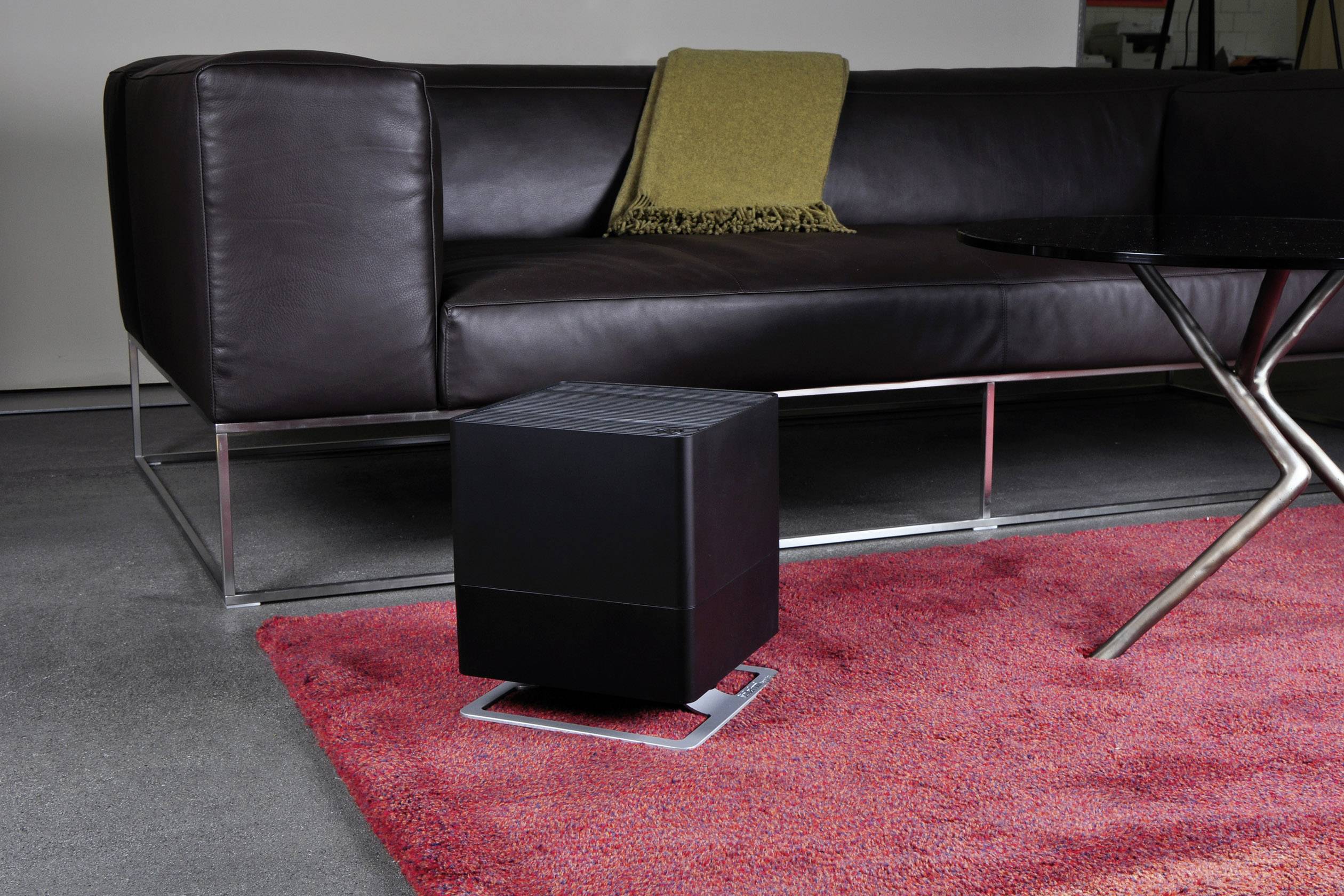 A black, cube-shaped piece of furniture on a red carpet, in front of a brown leather sofa with a yellow throw draped over its armrest.