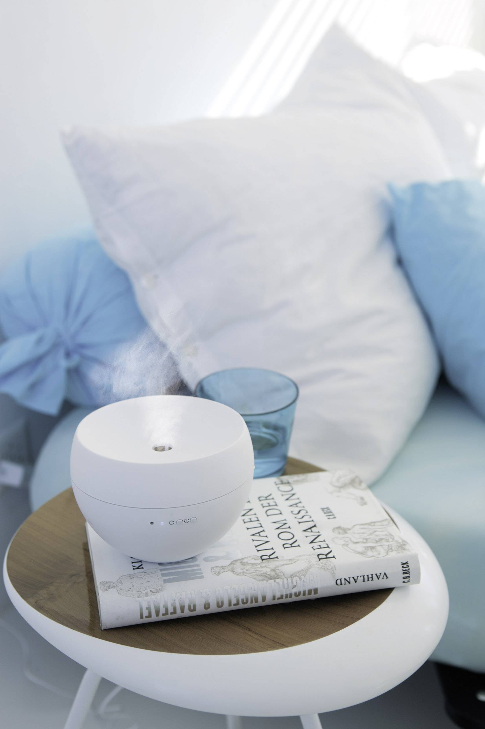 A humidifier stands on a small table next to a book titled 'Rivals Rally Adventure'. In the background, a sofa with cushions.