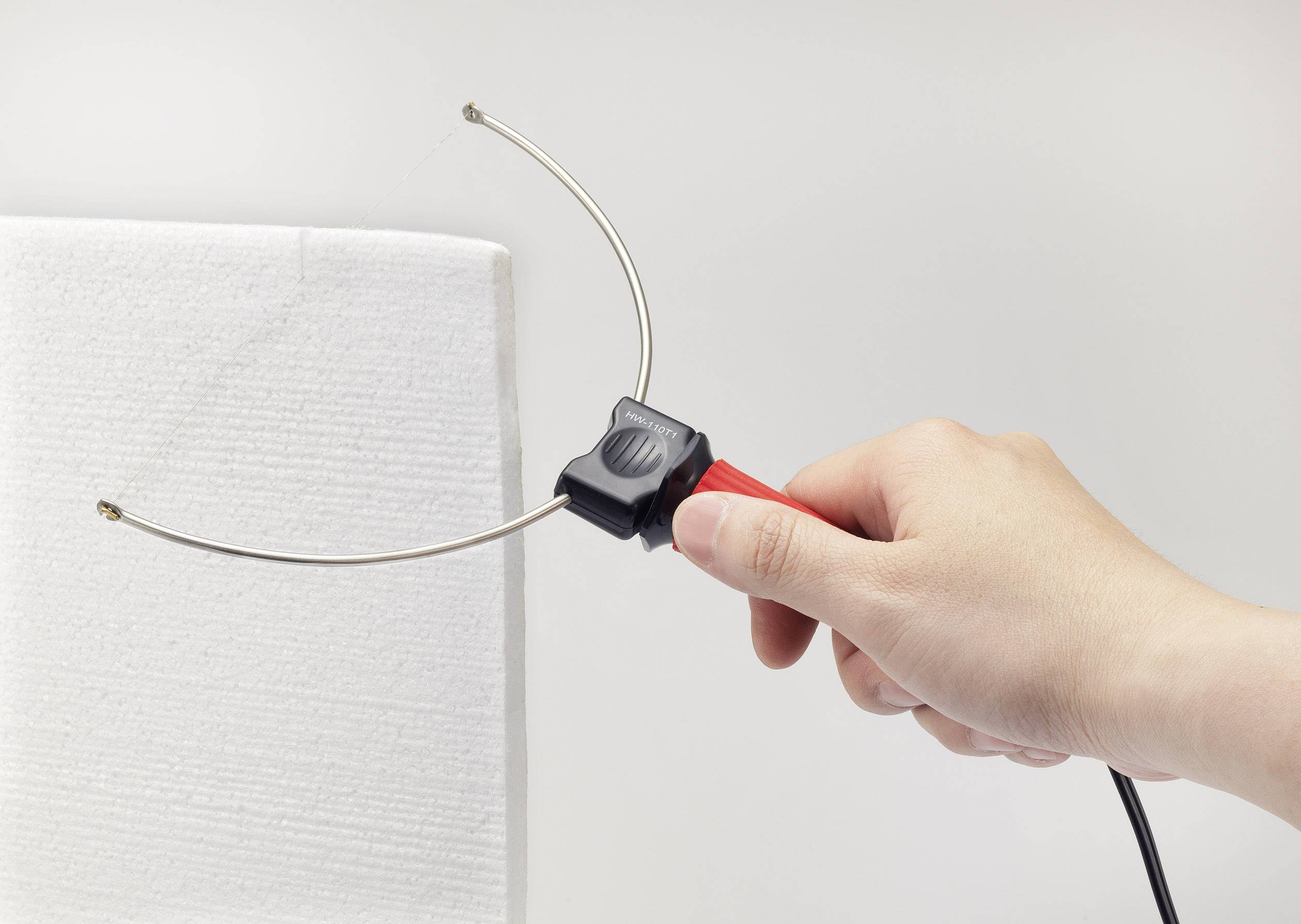A hand is holding a hot cutting tool that is cutting a polystyrene board. The tool has two curved metal rods.