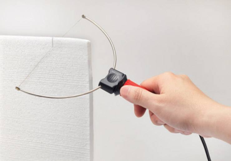 A hand holding an electric hot wire cutting tool is cutting into a white piece of foam.