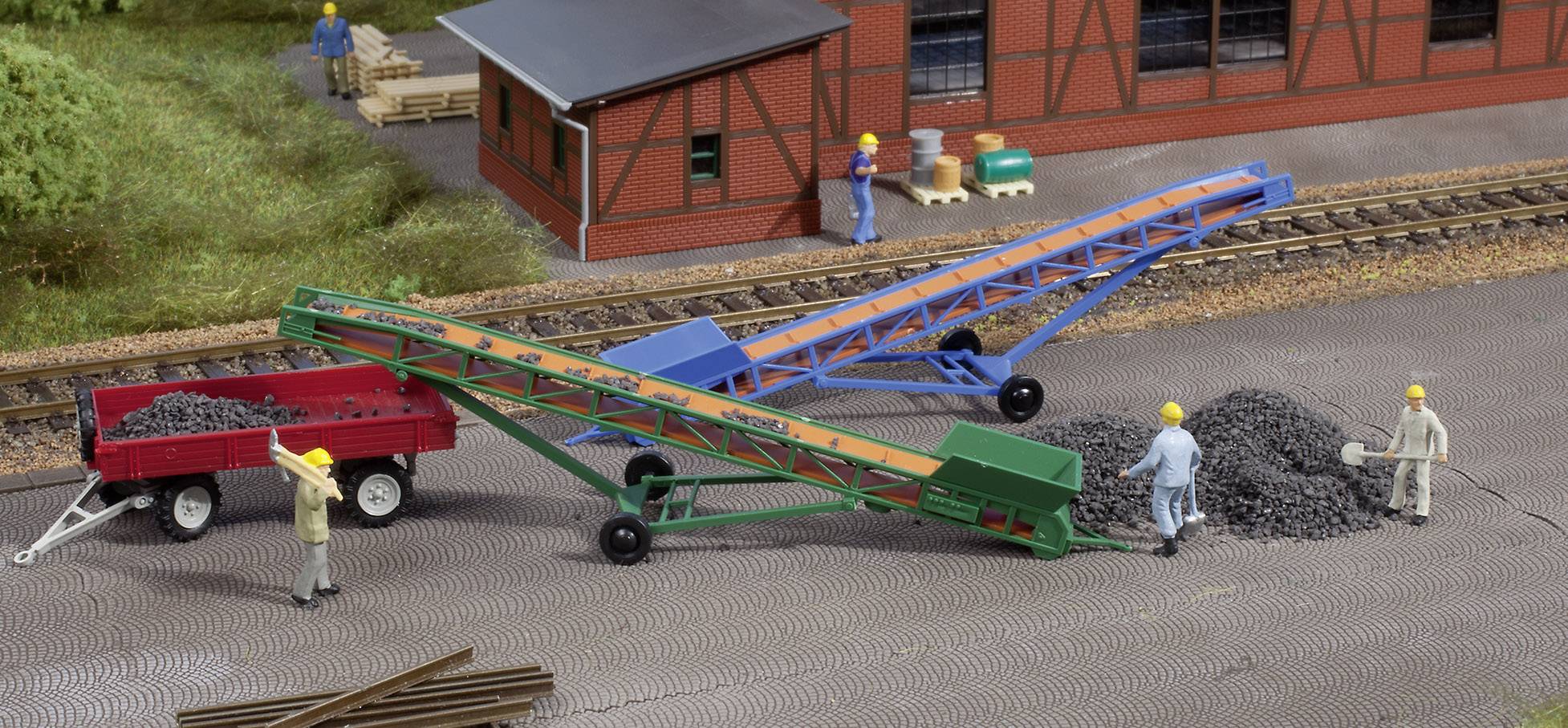 People are working on a model railway layout. Two conveyor belts and gravel piles can be seen, with workers standing in the background.