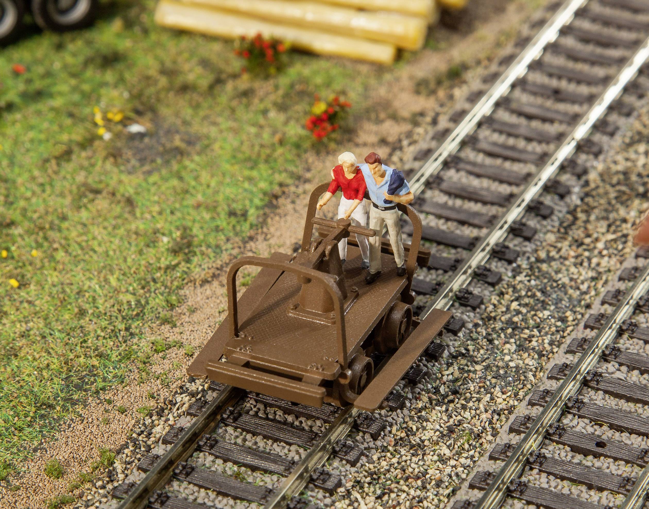 Two figures on a hand-operated rail trolley on a model railway track. Surroundings with grass and flowers.