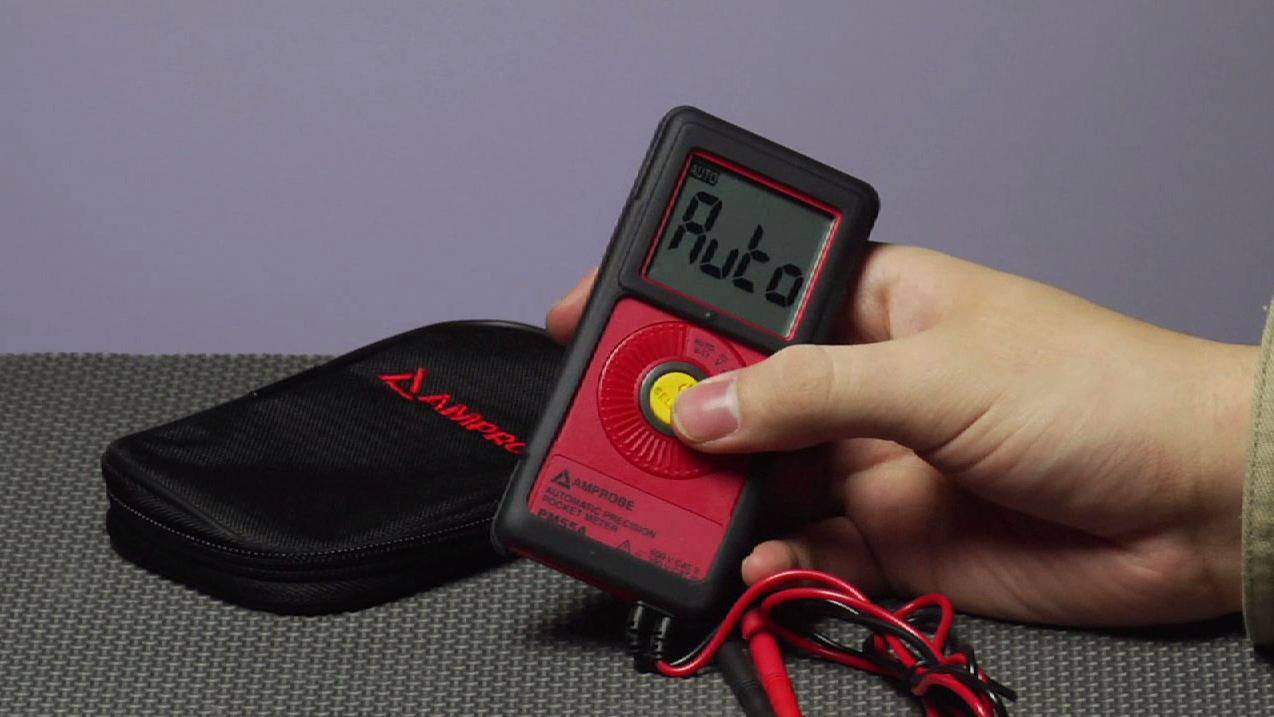 A person is holding a digital multimeter with an 'Auto' display, next to a black case. It is used for electrical measurements.