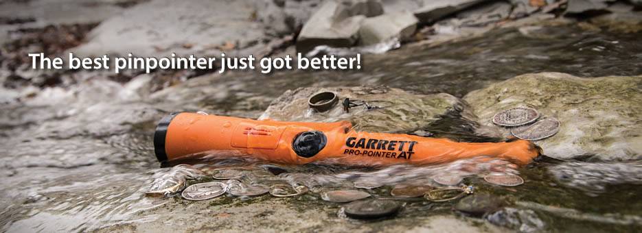 A orange metal detector lies in a shallow body of water on stones, surrounded by coins. Text: 'The best pinpointer just got better!'