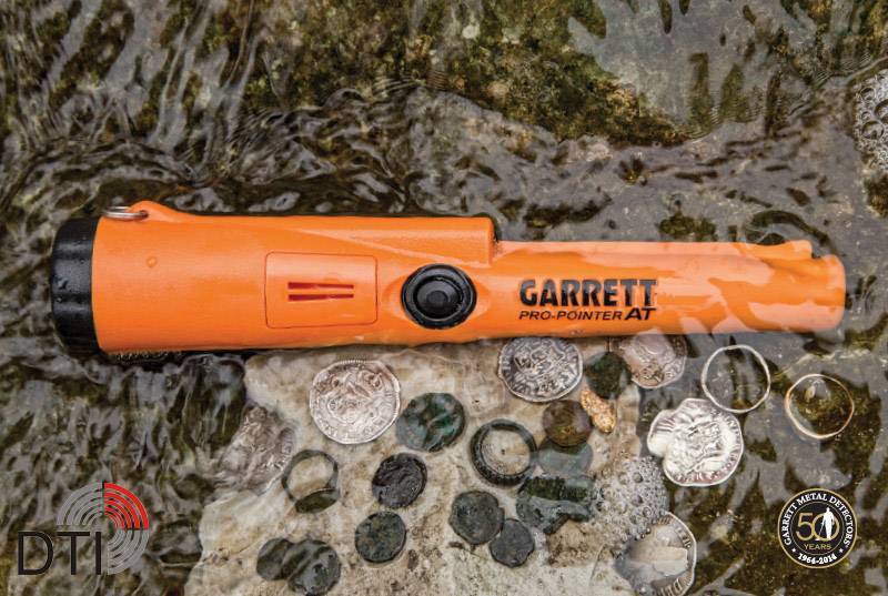 Orange metal detector lies on wet gravel amongst various coins and rings. Logos for DTI and a 50th anniversary are visible.