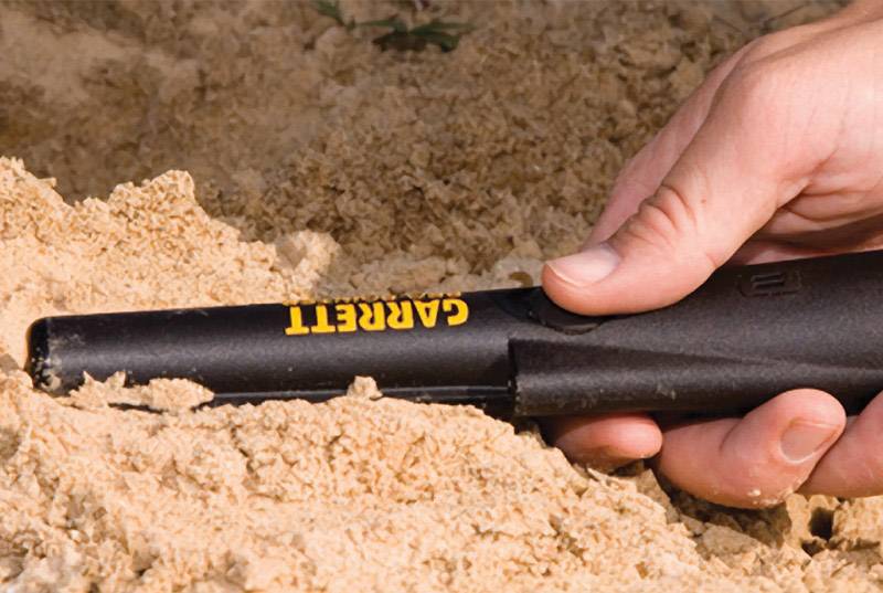 A hand holds a metal detector, partially buried in the sand. The device is black with a yellow 'Garrett' inscription.