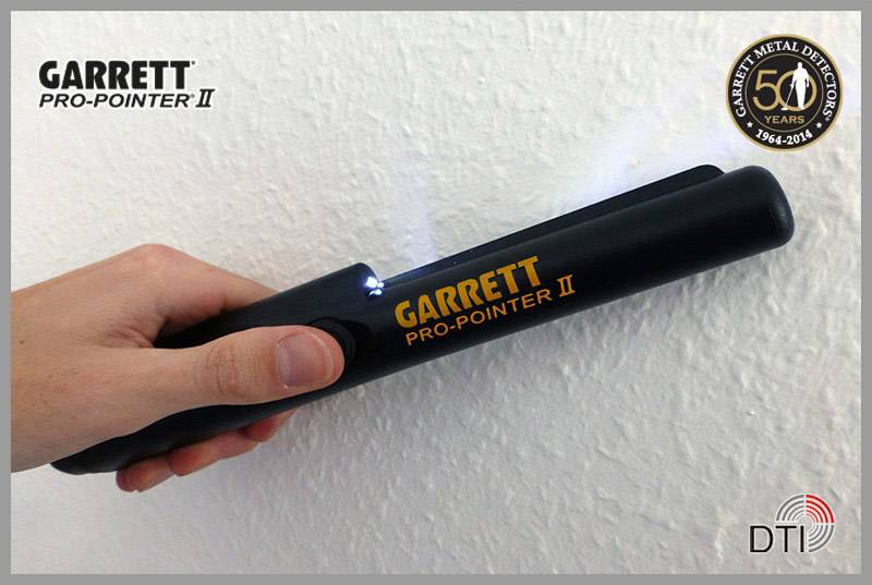 A hand is holding a black metal detector named 'Garrett Pro-Pointer II', which is being used with a light against a white wall.