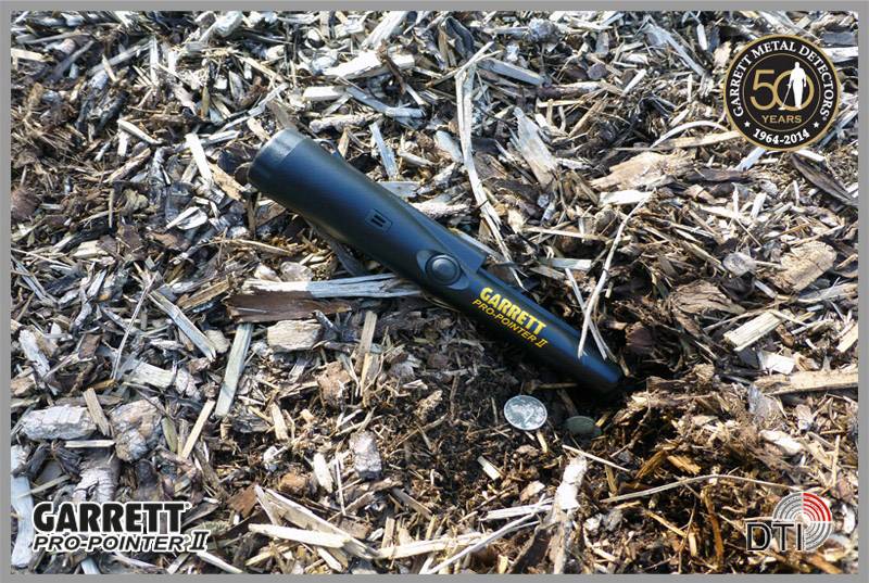 A black metal detector from the brand 'Garrett Pro-Pointer II' is lying on a wooden chip floor. A coin is next to it.