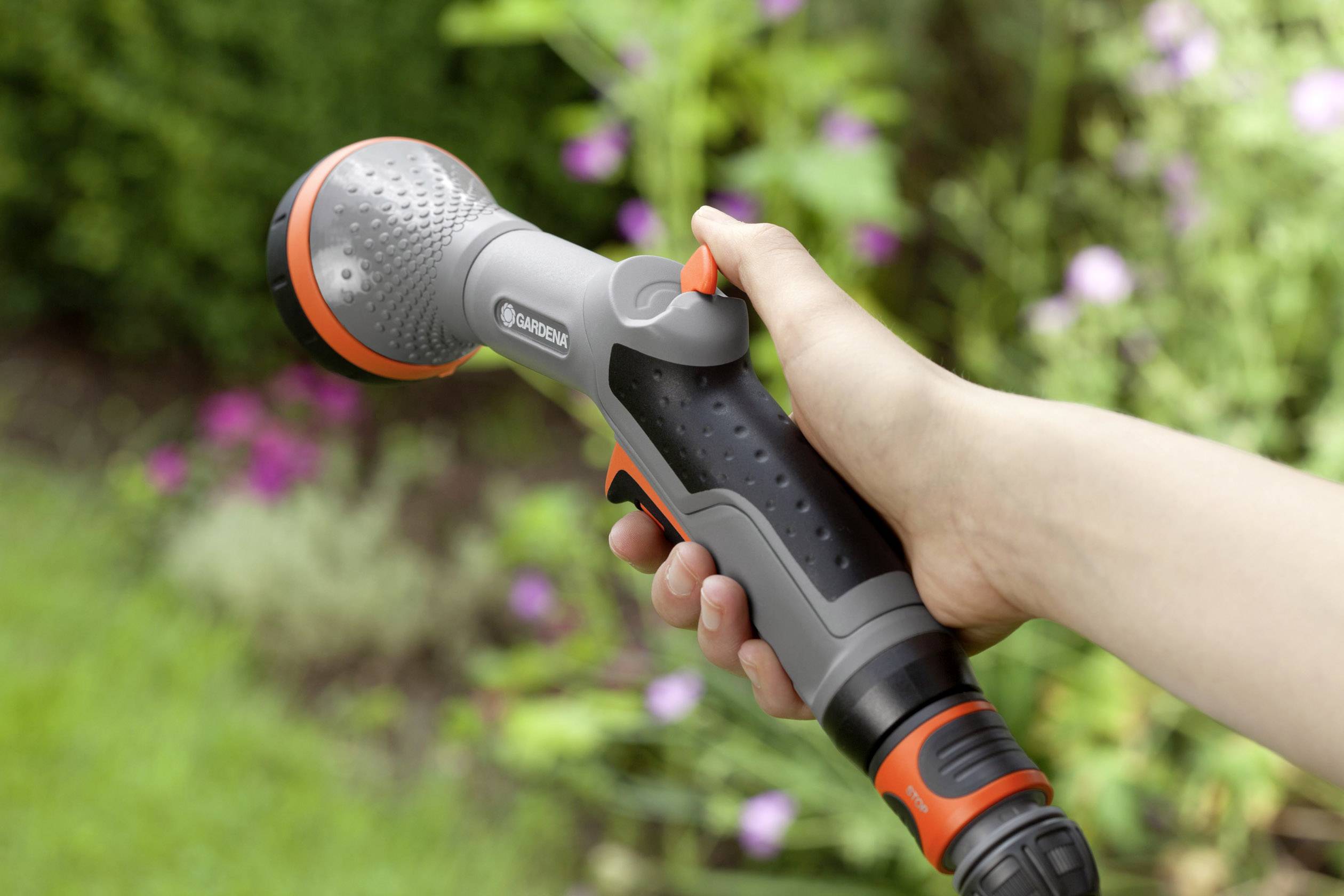A hand is holding a grey and orange garden spray gun with an adjustable water nozzle head, against a blurred garden background.