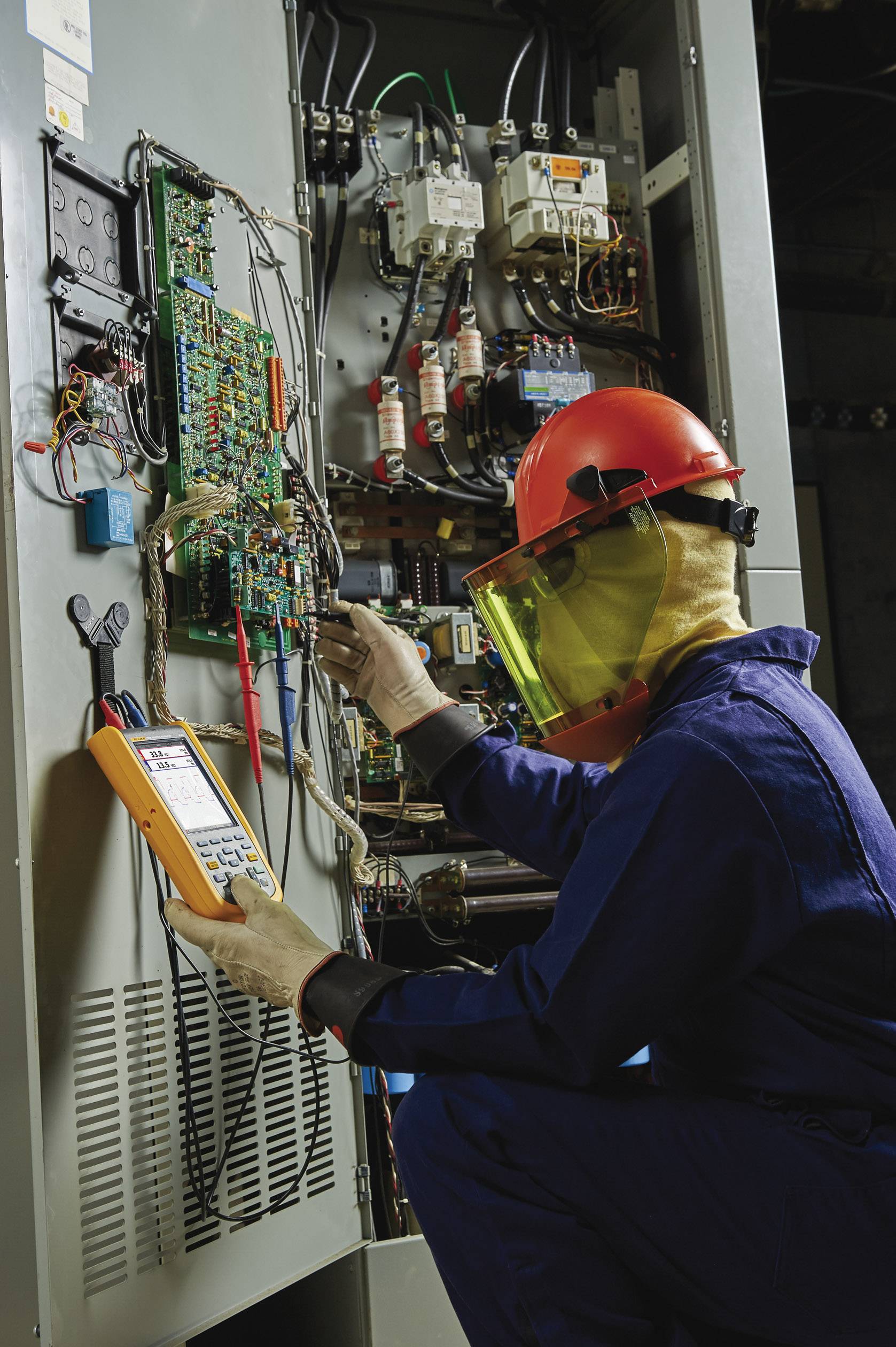 An electrician in protective clothing is working with a multimeter on an open electrical cabinet full of cables and fuses.