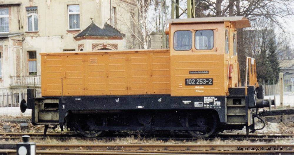 An orange locomotive stands on a track in front of old buildings. Inscription: 'Track Construction Vehicle' and '102 253-2'.